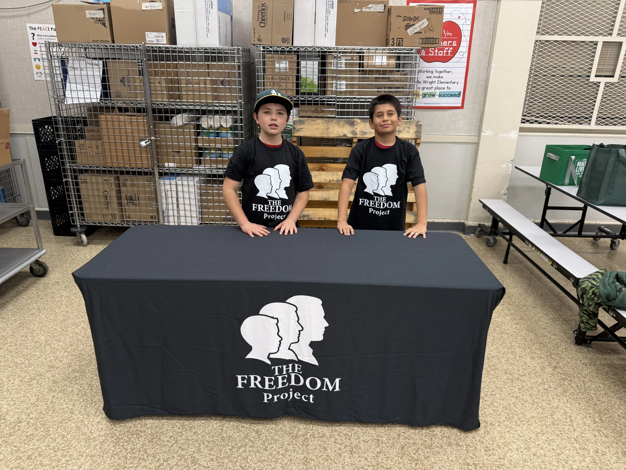 Two boys wearing black T-shirts with 'The Freedom Project' logo standing behind a table with the same logo on the tablecloth in a storage room.