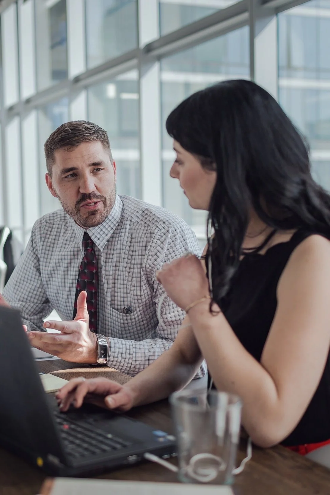 Two coworkers engaged in a discussion in a modern office with large windows, one man gesturing as he speaks, and a woman listening and working on a laptop.