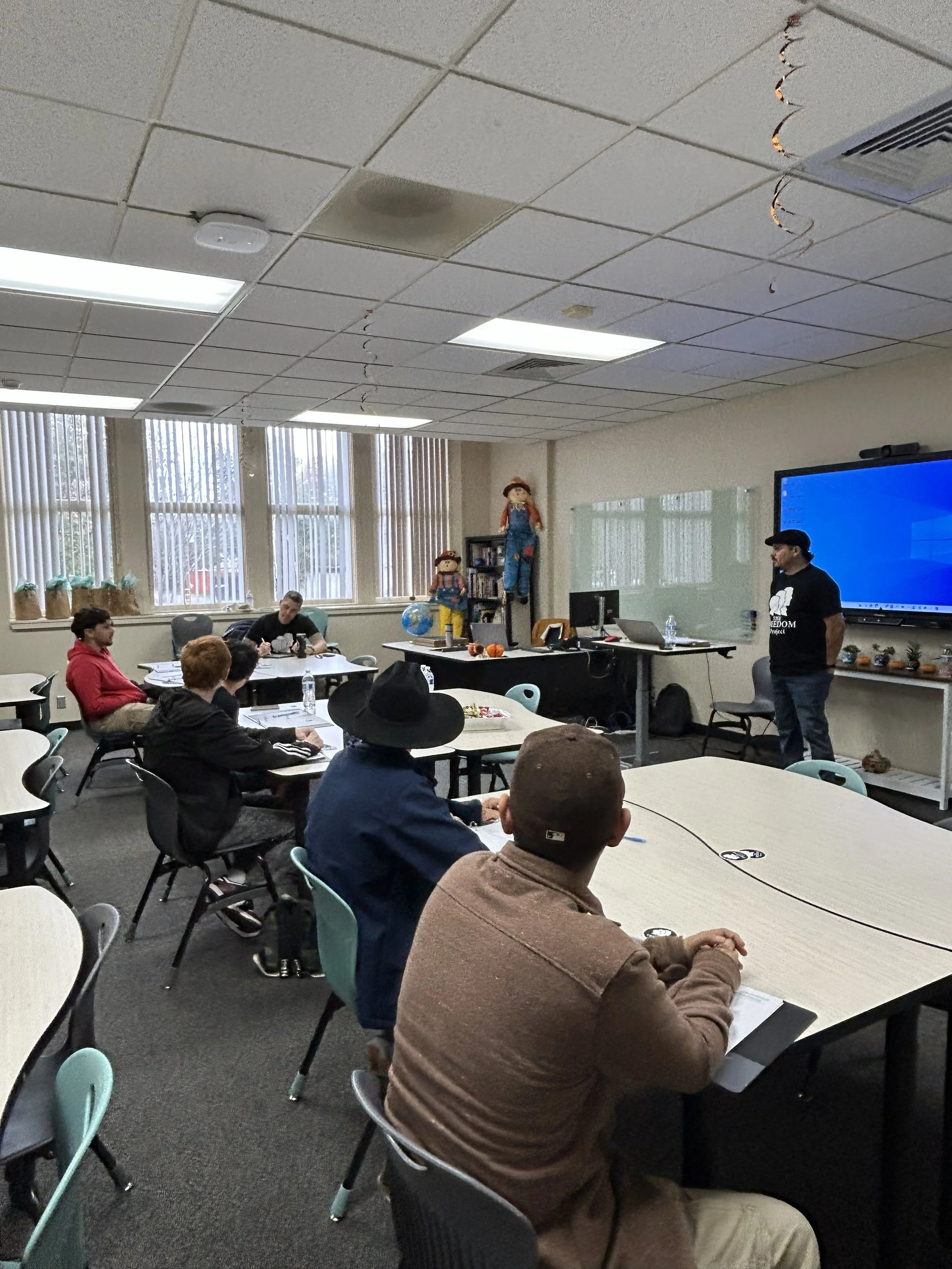 A classroom or meeting room with several people seated at tables, listening to a person standing at the front near a large screen. Decorations include dolls and plants on the shelves, and there are windows with vertical blinds letting in natural light.
