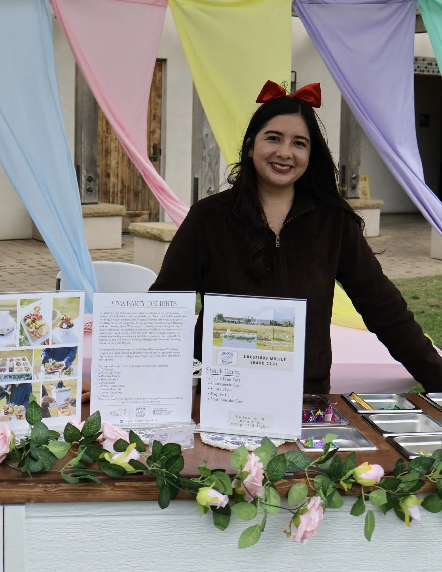 Woman standing at a decorated booth with colorful drapes and a display of informational flyers. The booth has floral garlands and small trays in front. She is smiling and wearing a red bow in her hair.