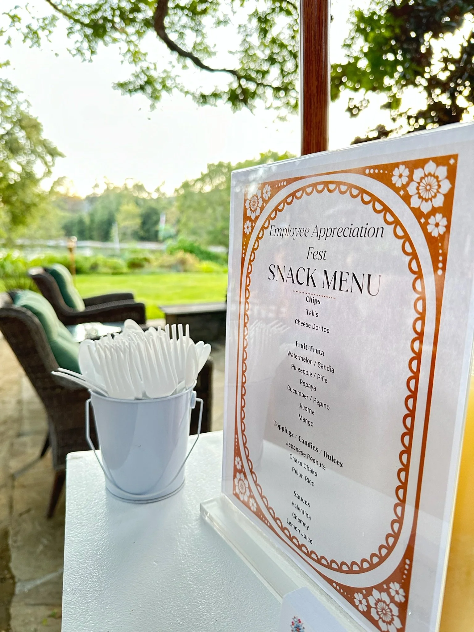 Outdoor setting with a snack menu displayed on a stand and a small bucket of white plastic forks. The menu is titled "Employee Appreciation Fest Snack Menu" and lists various snacks and sauces. There are outdoor chairs and greenery in the background.