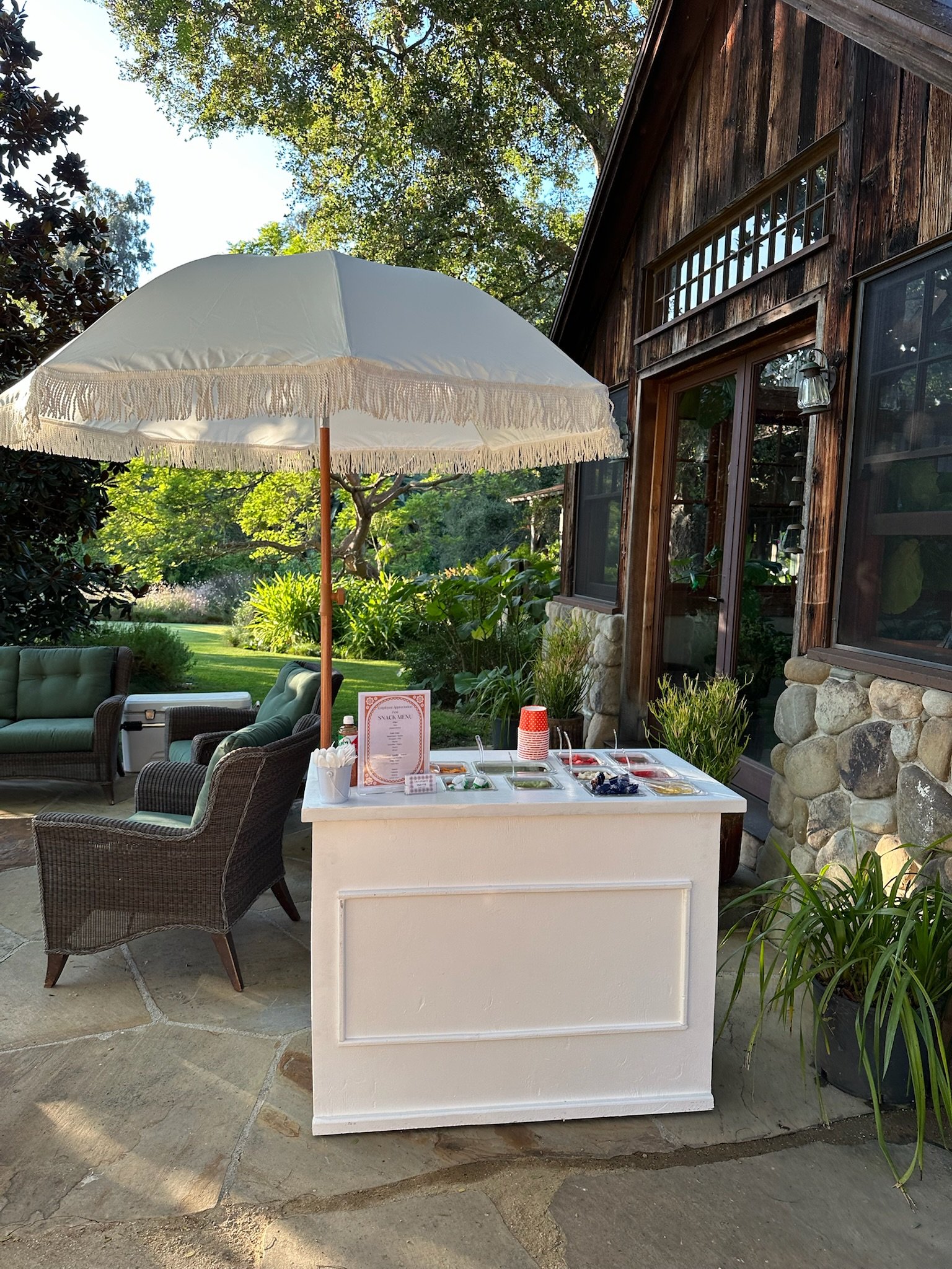 Outdoor ice cream cart with umbrella, surrounded by wicker chairs, greenery, and a stone building.