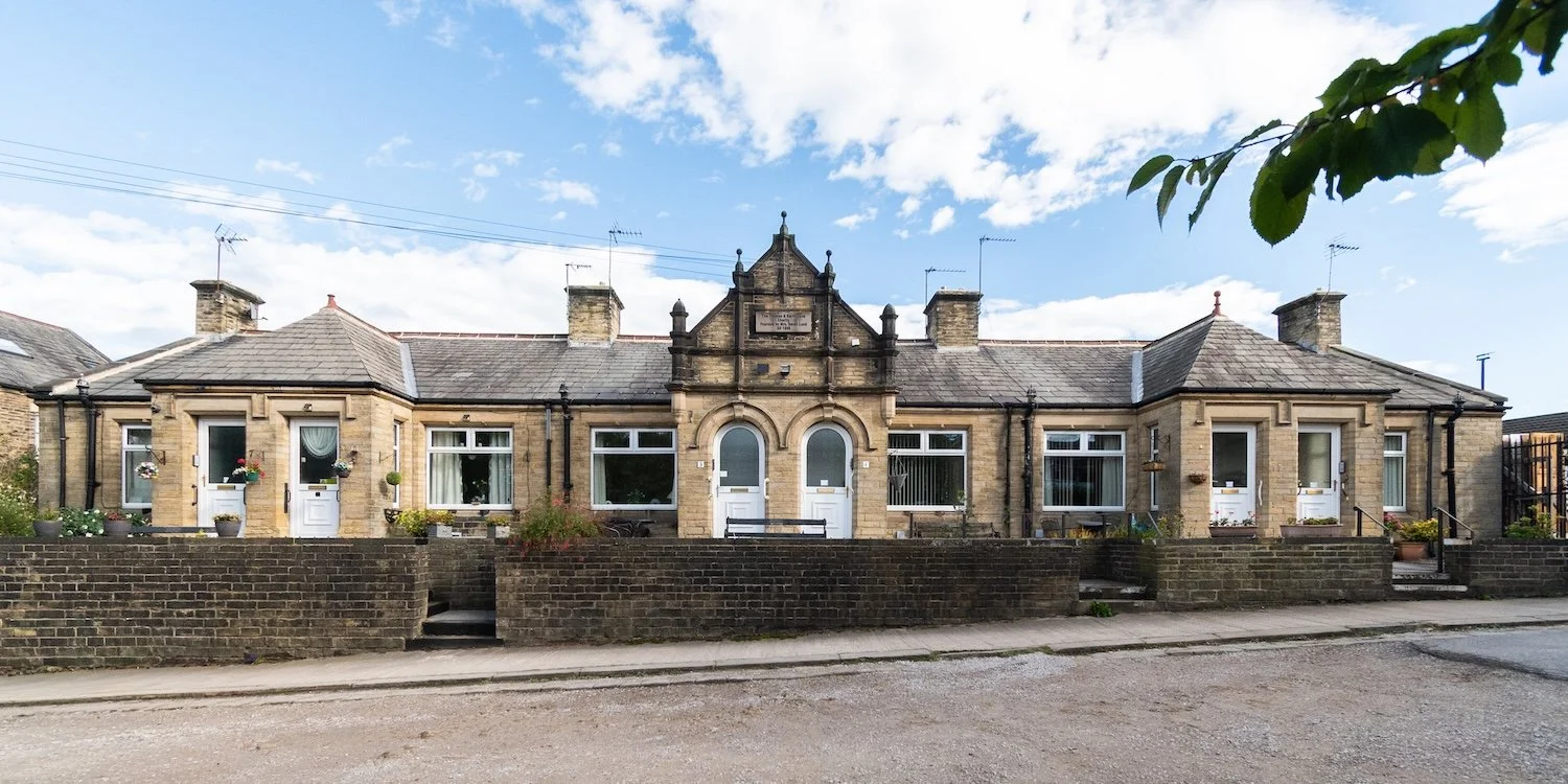 A single-story stone building with arched windows and a central decorative gable, surrounded by a low brick wall and a sidewalk, under a partly cloudy sky.