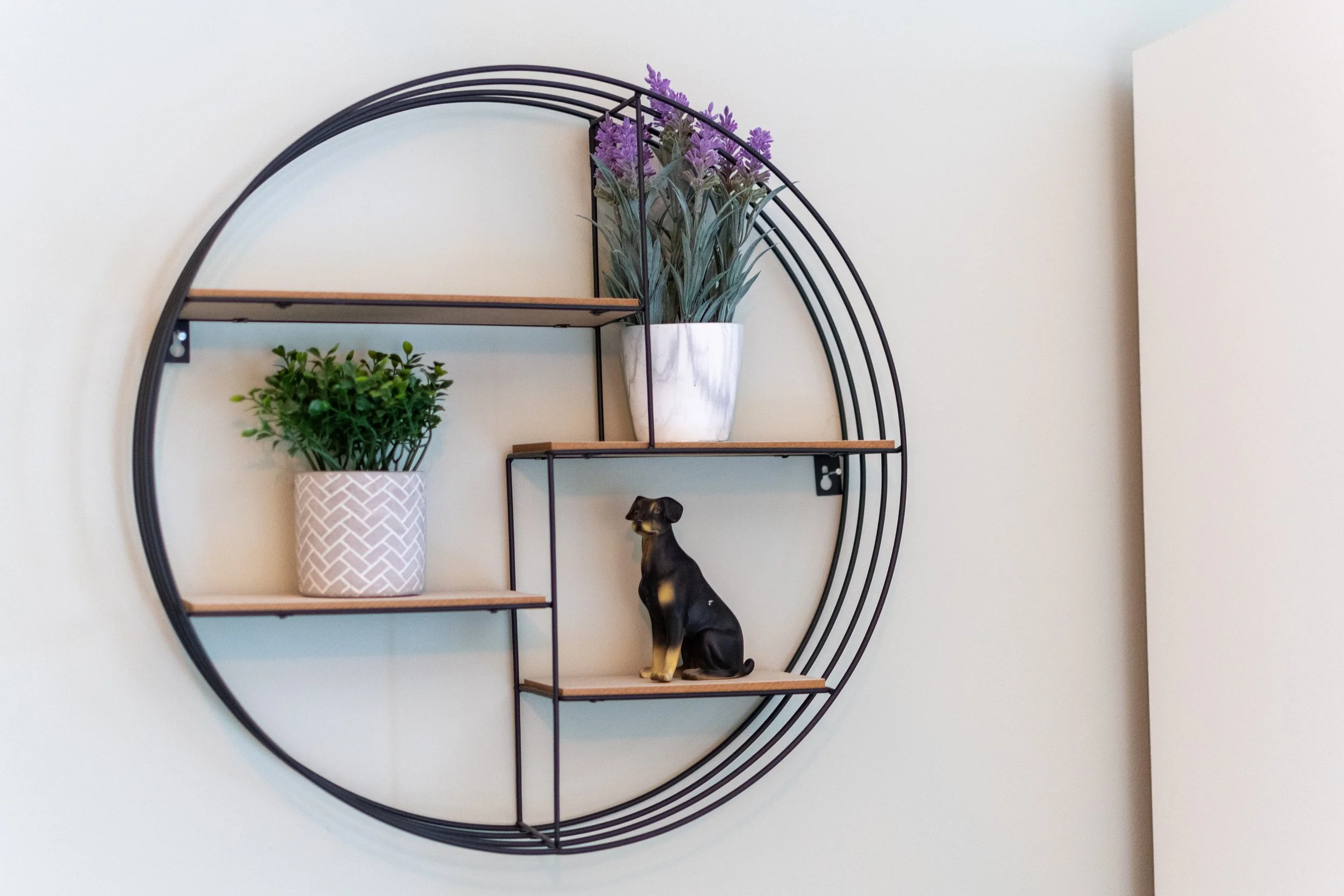 A circular wall shelf with black metal frame and wooden shelves, decorated with potted plants and a dog statue, against a plain white wall.