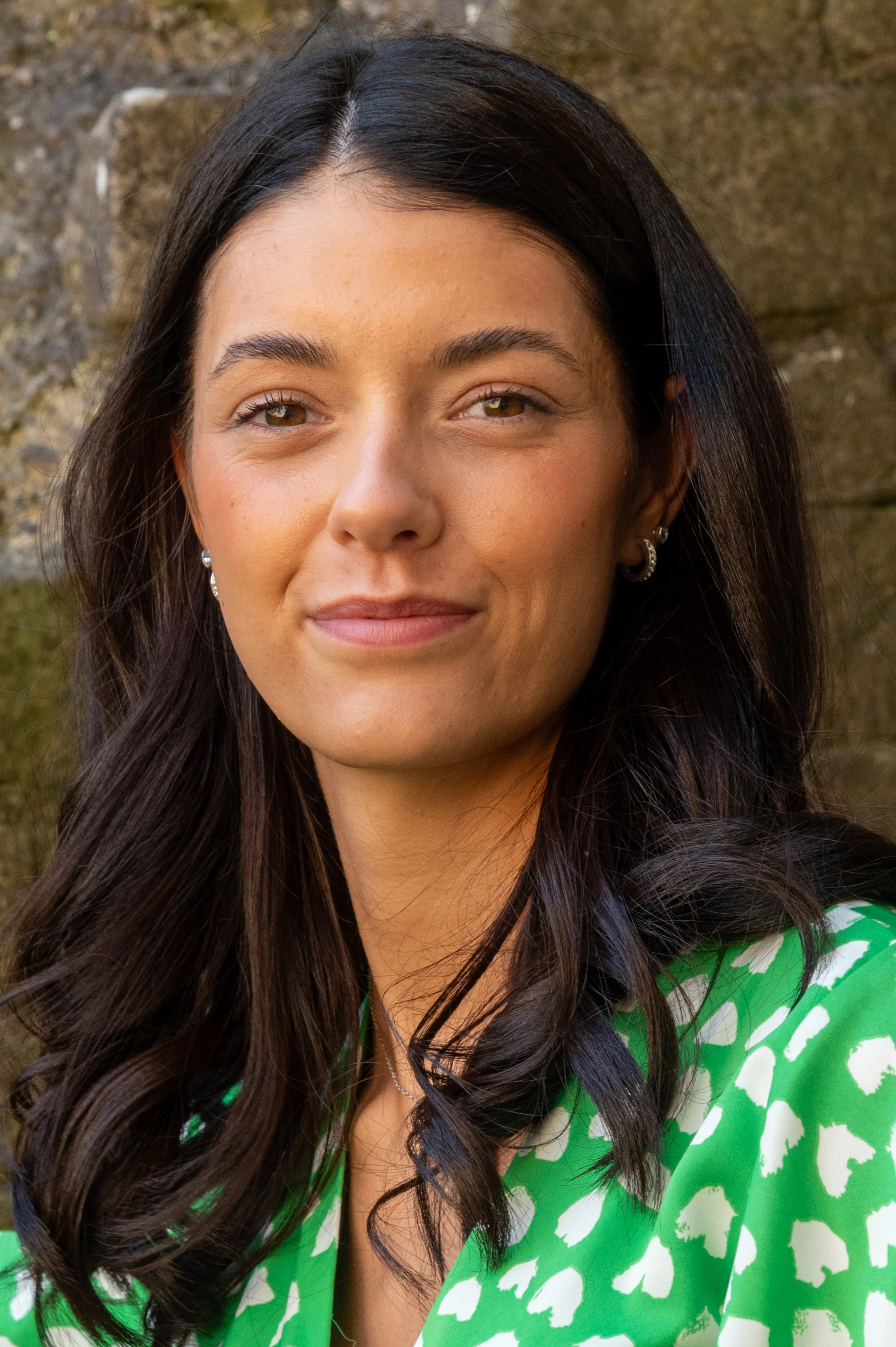 Close-up of a young woman with long dark hair wearing a green and white patterned top, standing outdoors with a stone wall in the background.