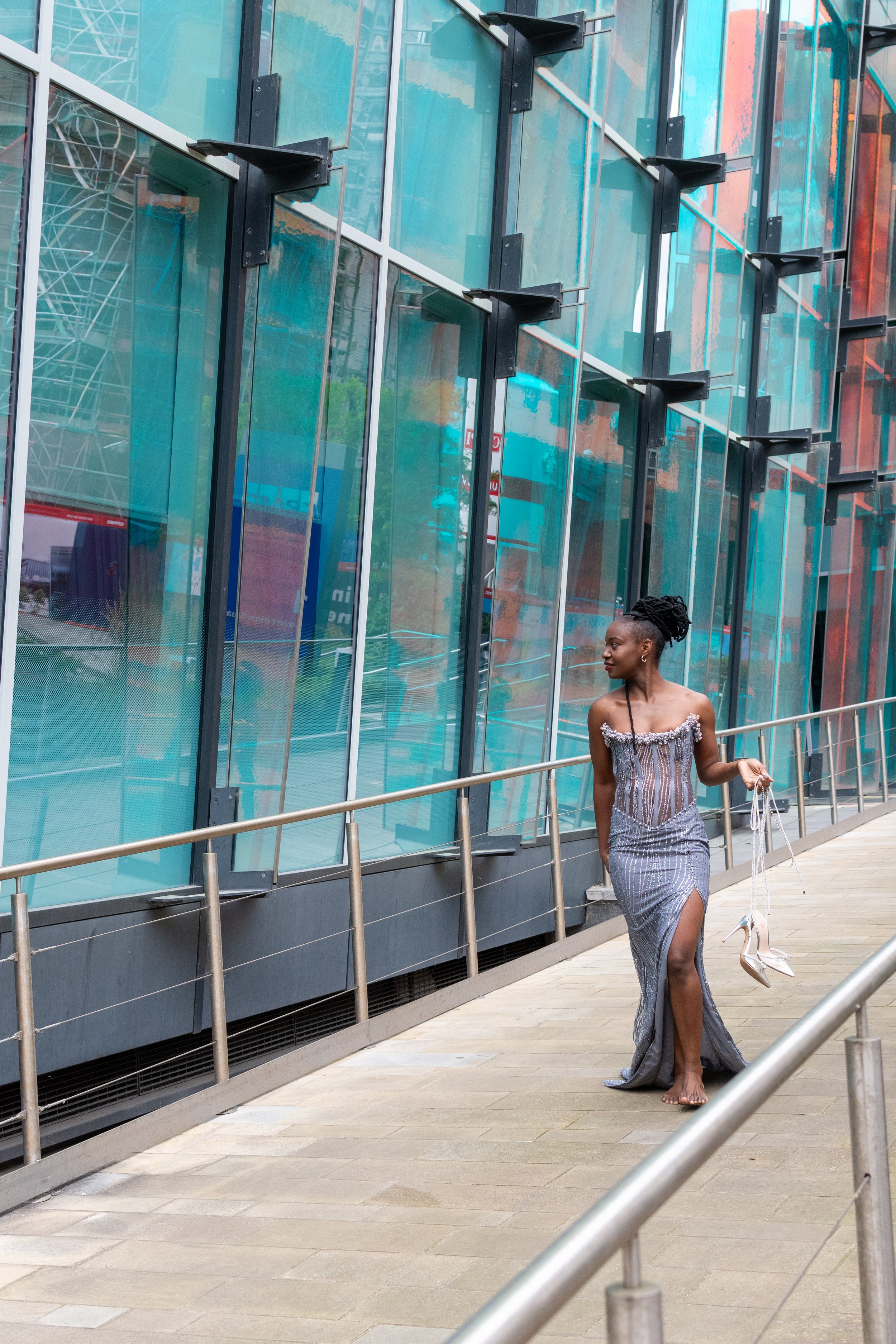 A woman dressed in an elegant, silver, strapless evening gown walks barefoot outdoors by a modern building with glass panels. She is holding a pair of silver high-heeled shoes in her right hand.