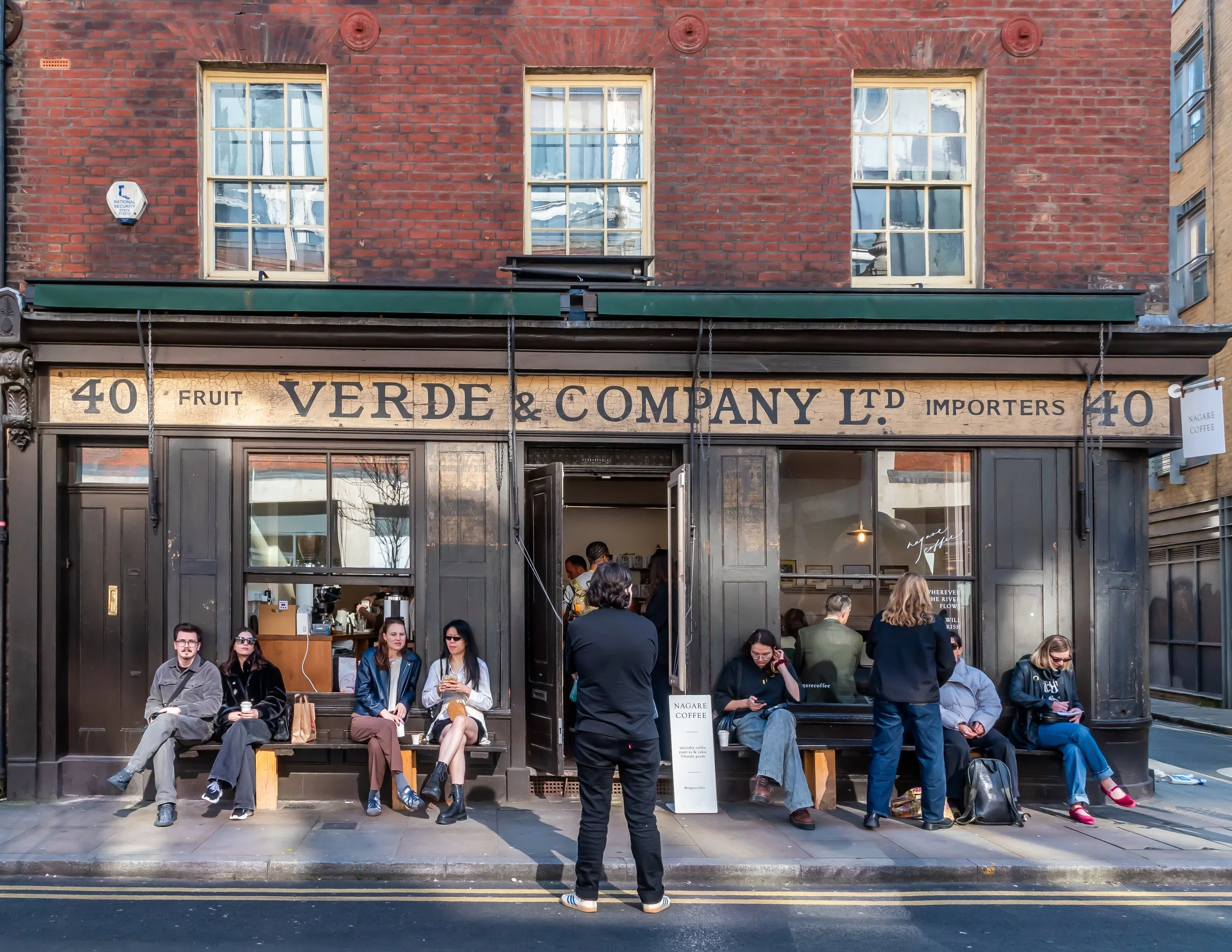 People sitting and standing outside a coffee shop named Verde & Company Ltd. The shop has a brick facade with three windows and an open door. Some individuals are chatting, while others are using their phones, and some are walking or sitting on benches.