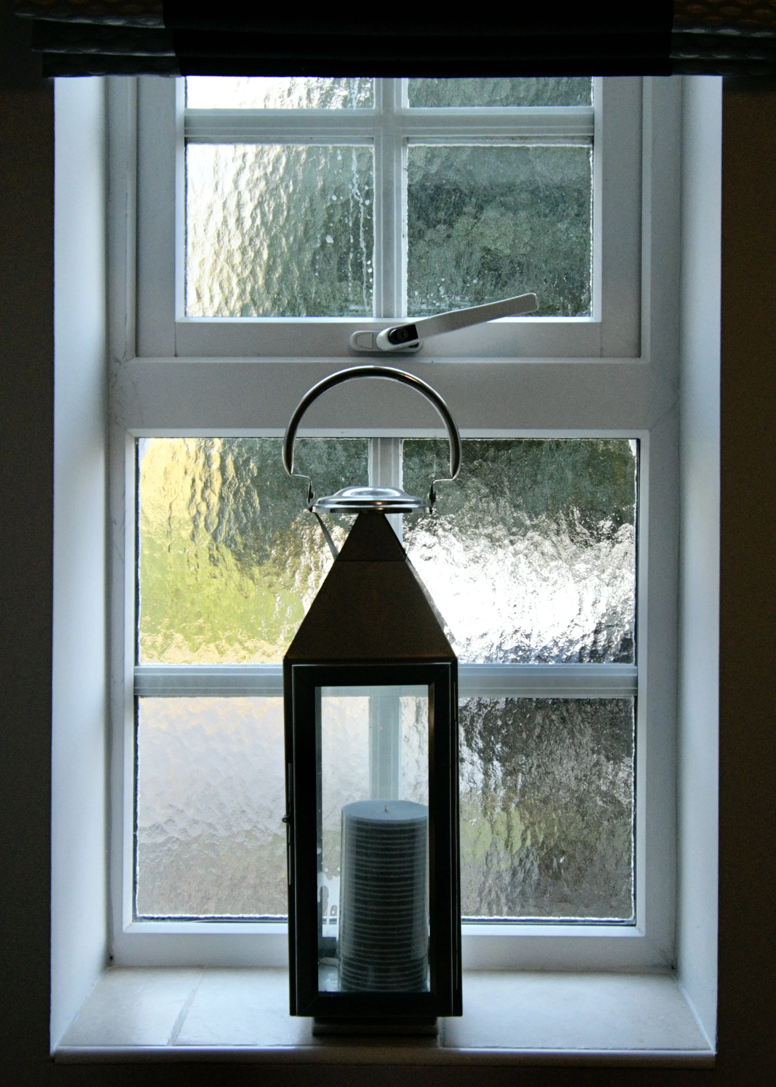 View through a window showing a water fountain with a metal arch and a circular base, with trees and greenery outside.