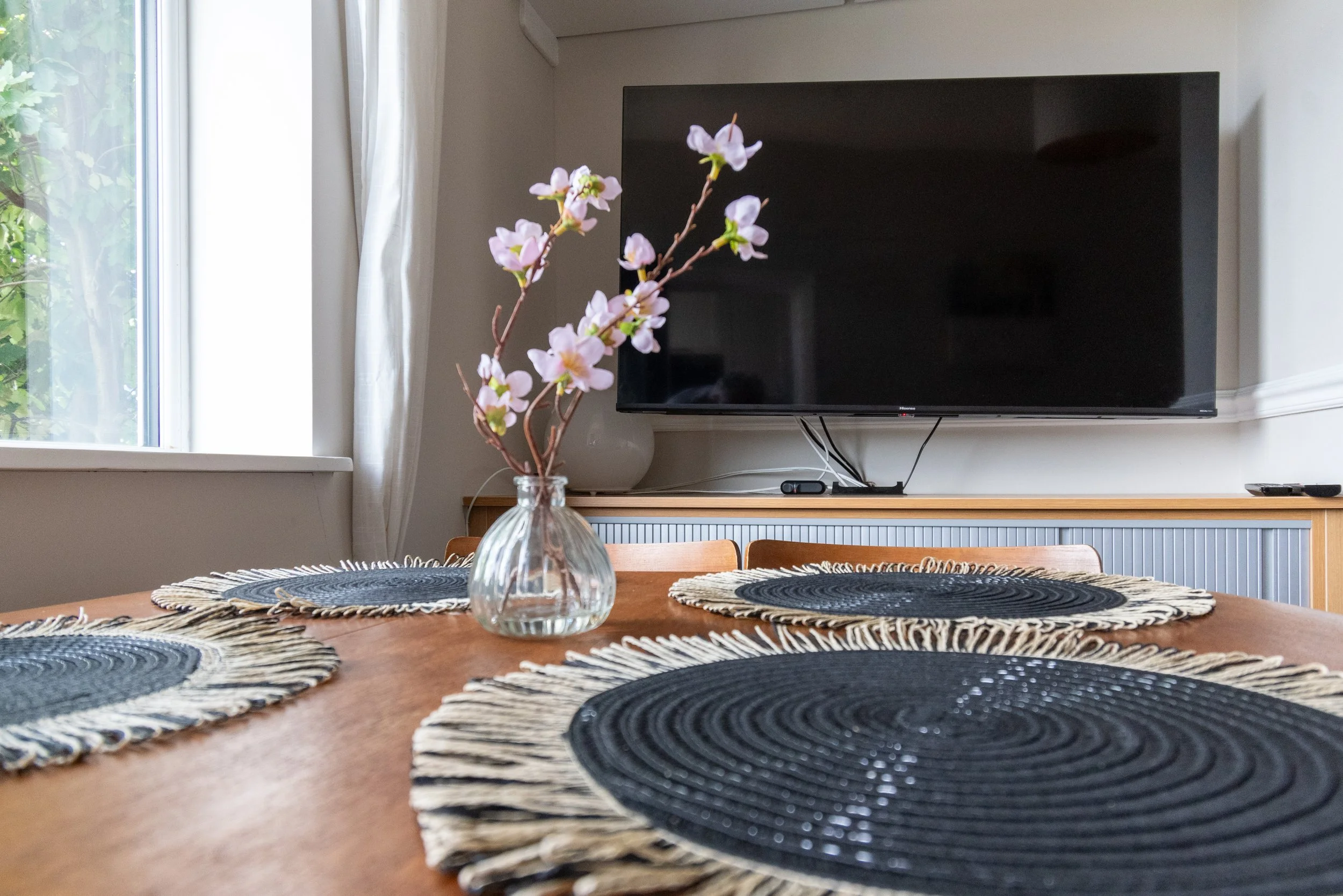 A dining room table with woven black and beige placemats, a glass vase with pink and white flowers, and a large flat-screen TV mounted on the wall in the background.