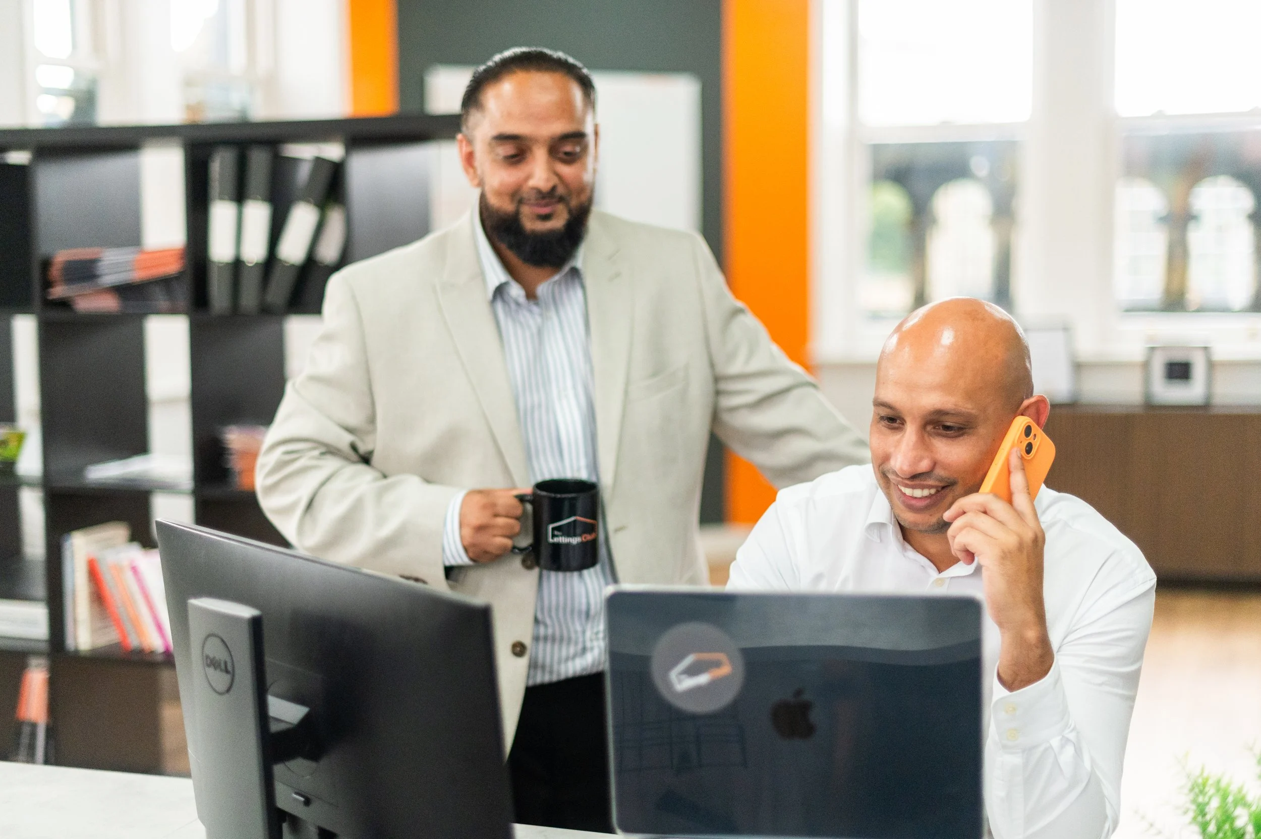 Two men working in an office, one smiling on the phone and the other holding a coffee mug, with computer monitors and bookshelves in the background.