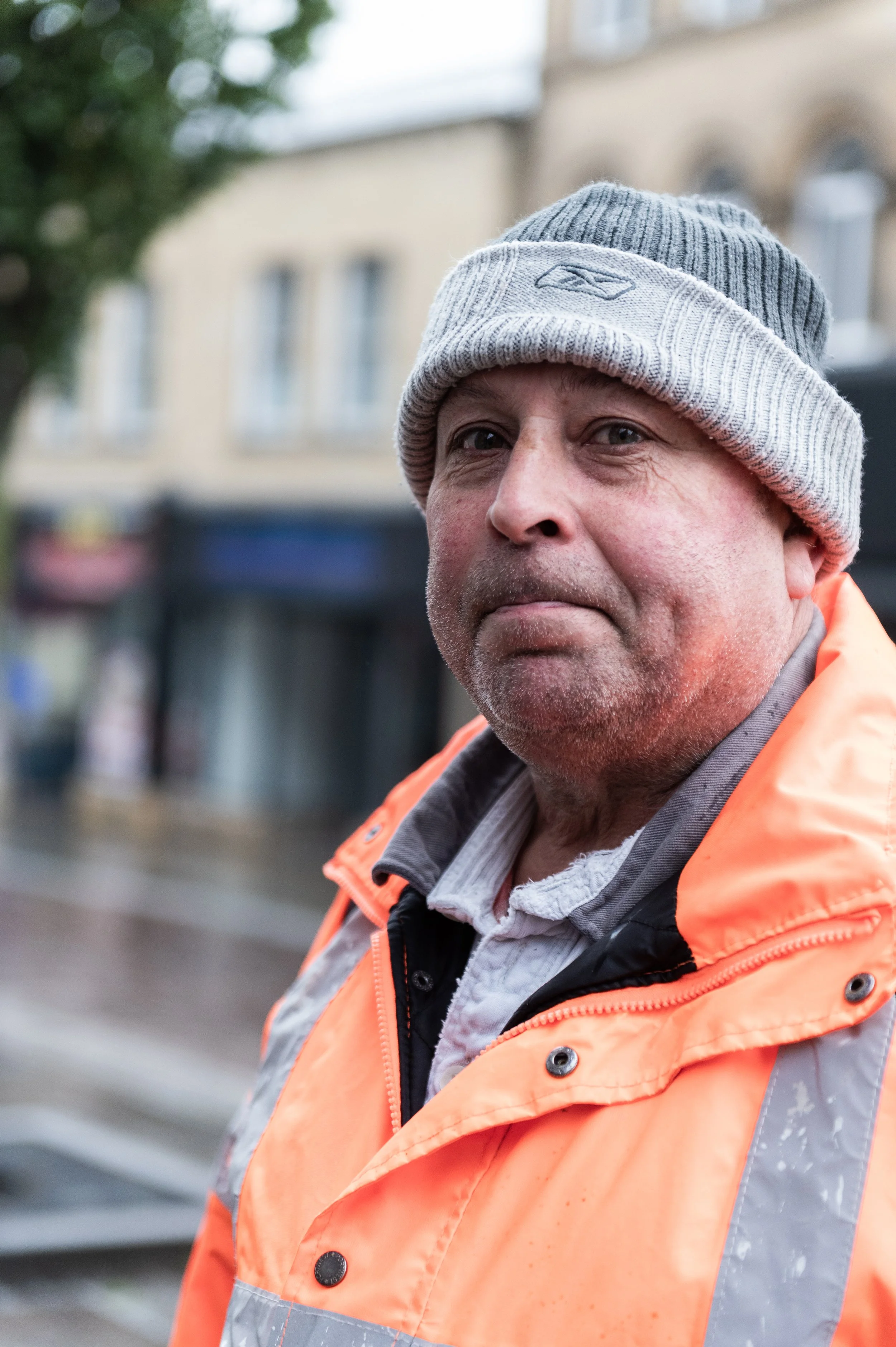 Close-up of a middle-aged man wearing a gray beanie and a high-visibility orange jacket, outdoors on a cloudy day.