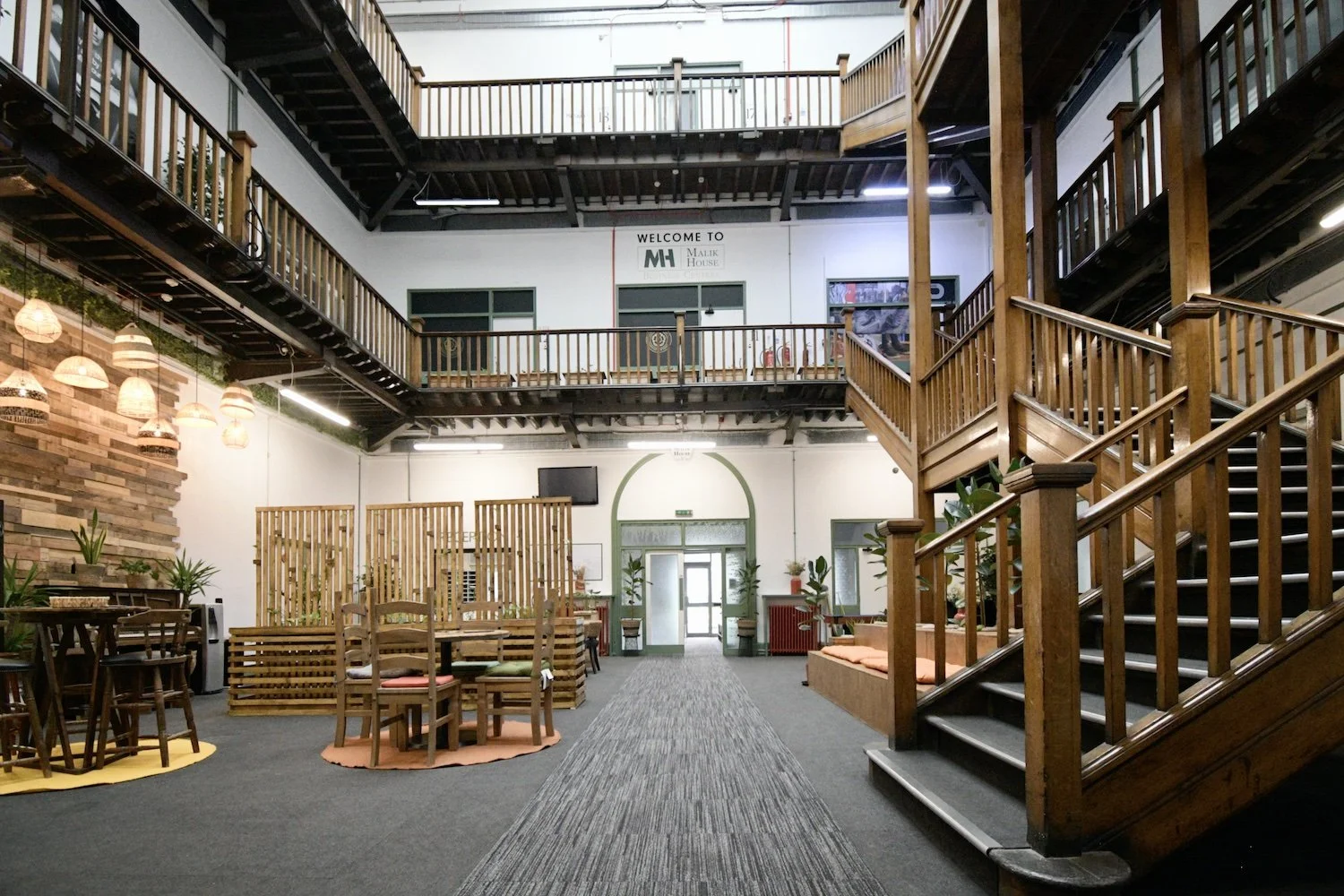 Interior of a large multi-level atrium or co-working space with wooden railings, staircase, decorative lighting, and plants.