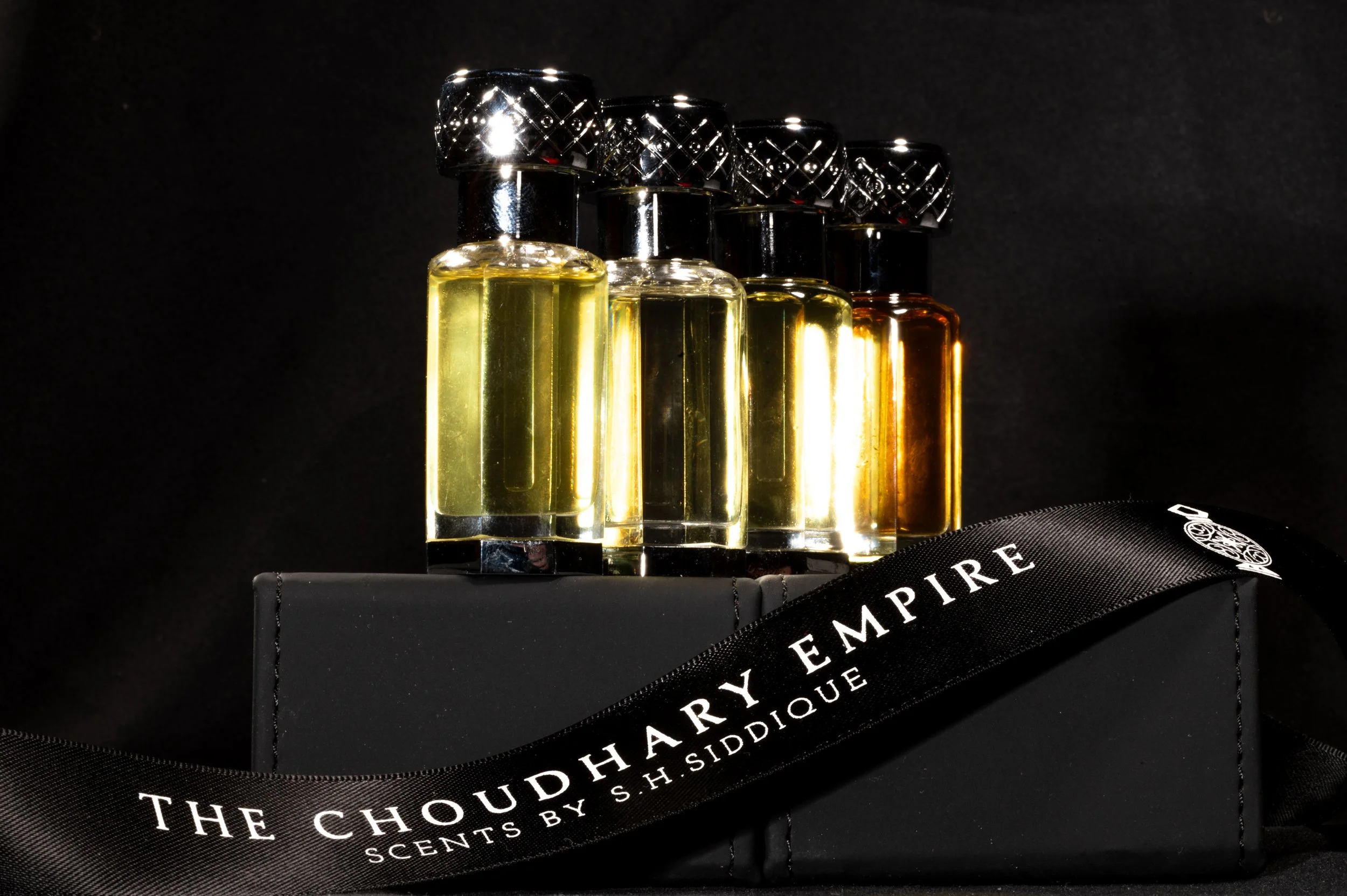 A set of five perfume bottles with black caps and golden liquid, arranged in a row on a black surface against a dark background, with a black box and company ribbon.