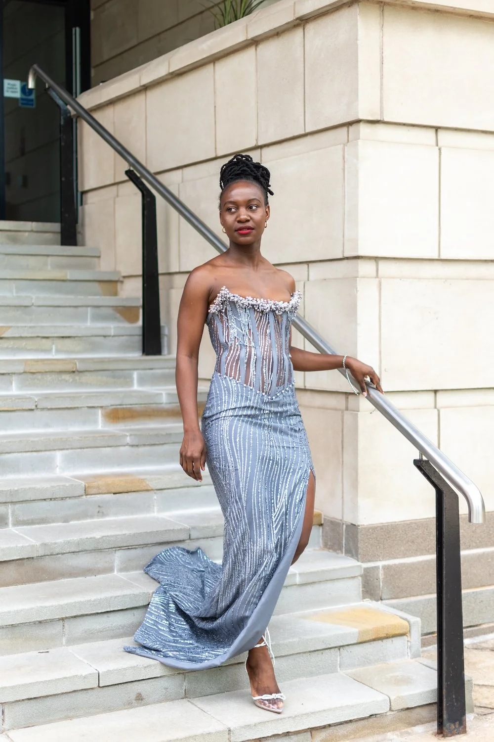 A woman in an elegant, strapless, silver gown with pearl embellishments, descending stairs outside a building with beige stone walls, holding a handrail, and looking to her left.