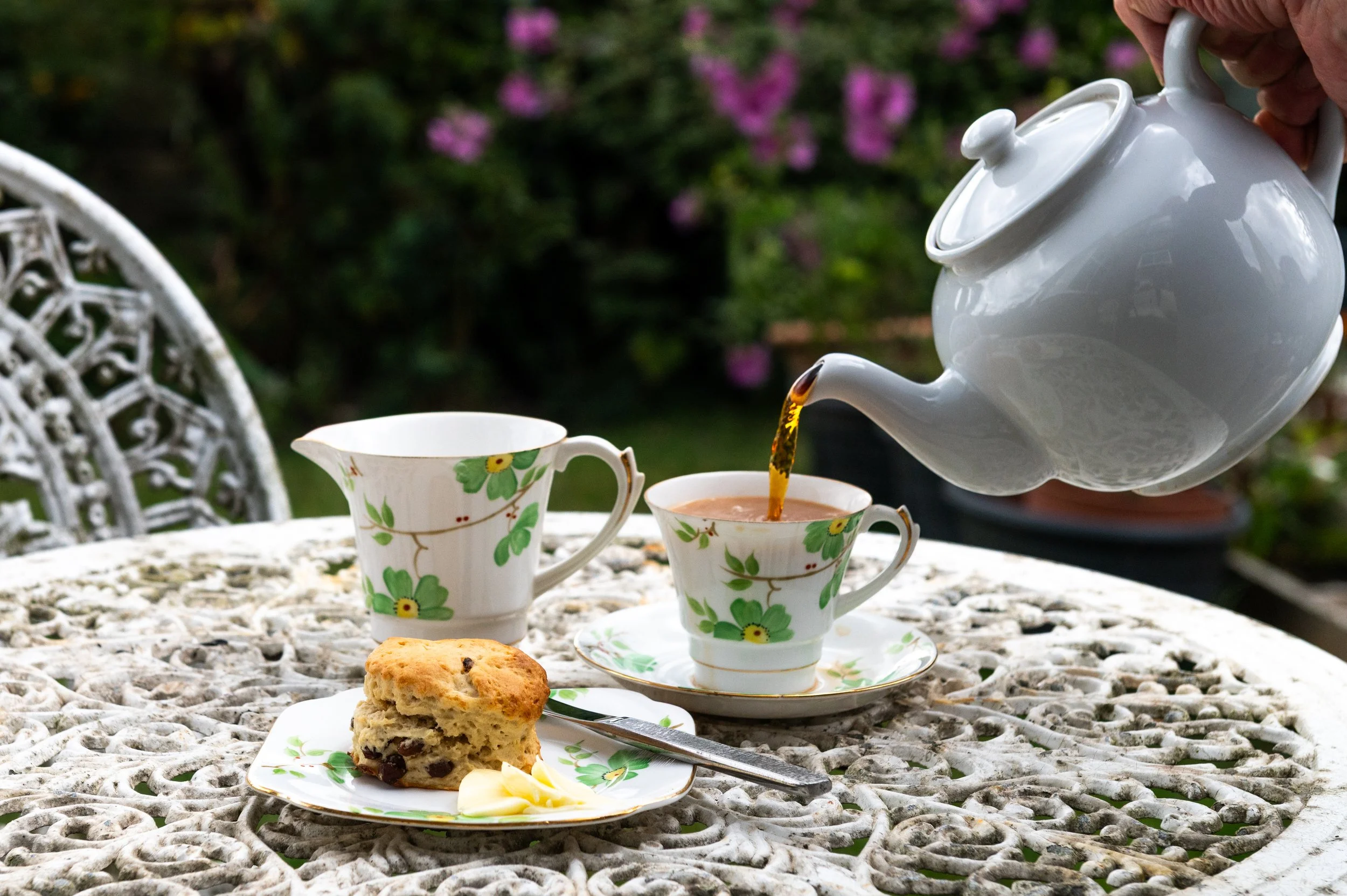A person pours tea from a white teapot into floral patterned teacups on a white lace table outdoors, with a cookie and butter on a plate in the foreground and a garden with pink flowers in the background.