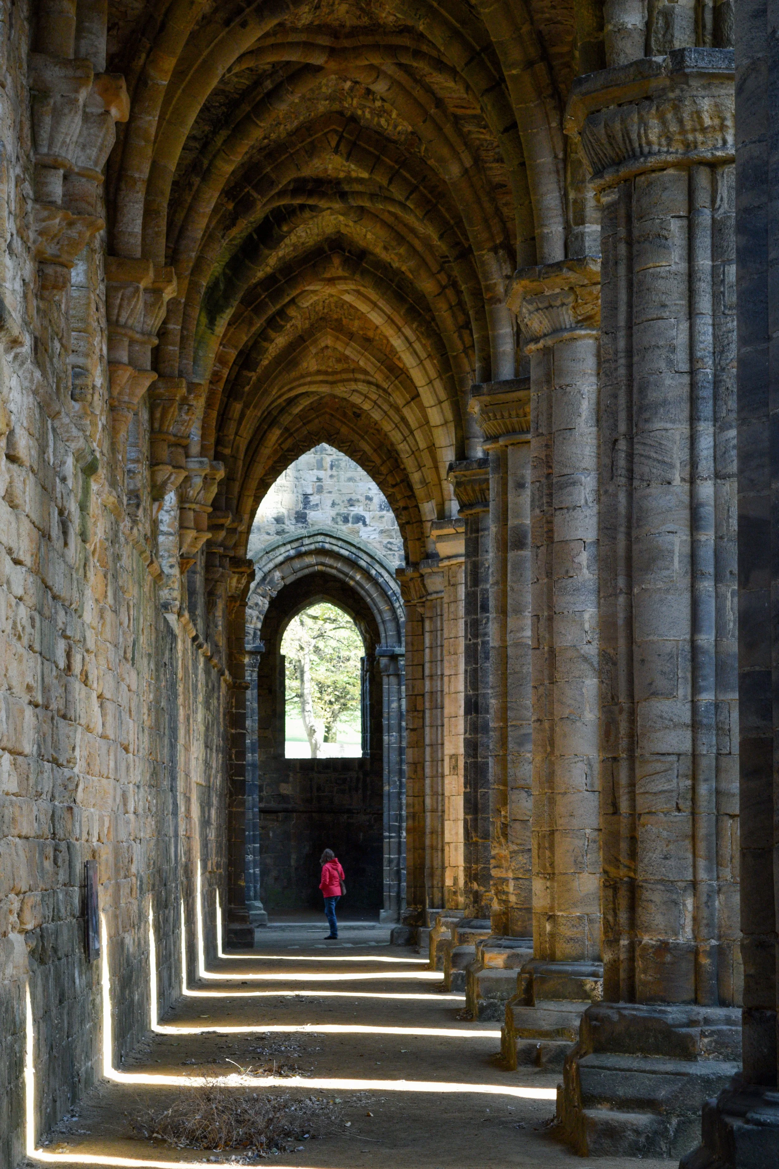 A stone corridor with arched ceilings and columns, with a person in a pink jacket walking through, illuminated by natural light from the end.