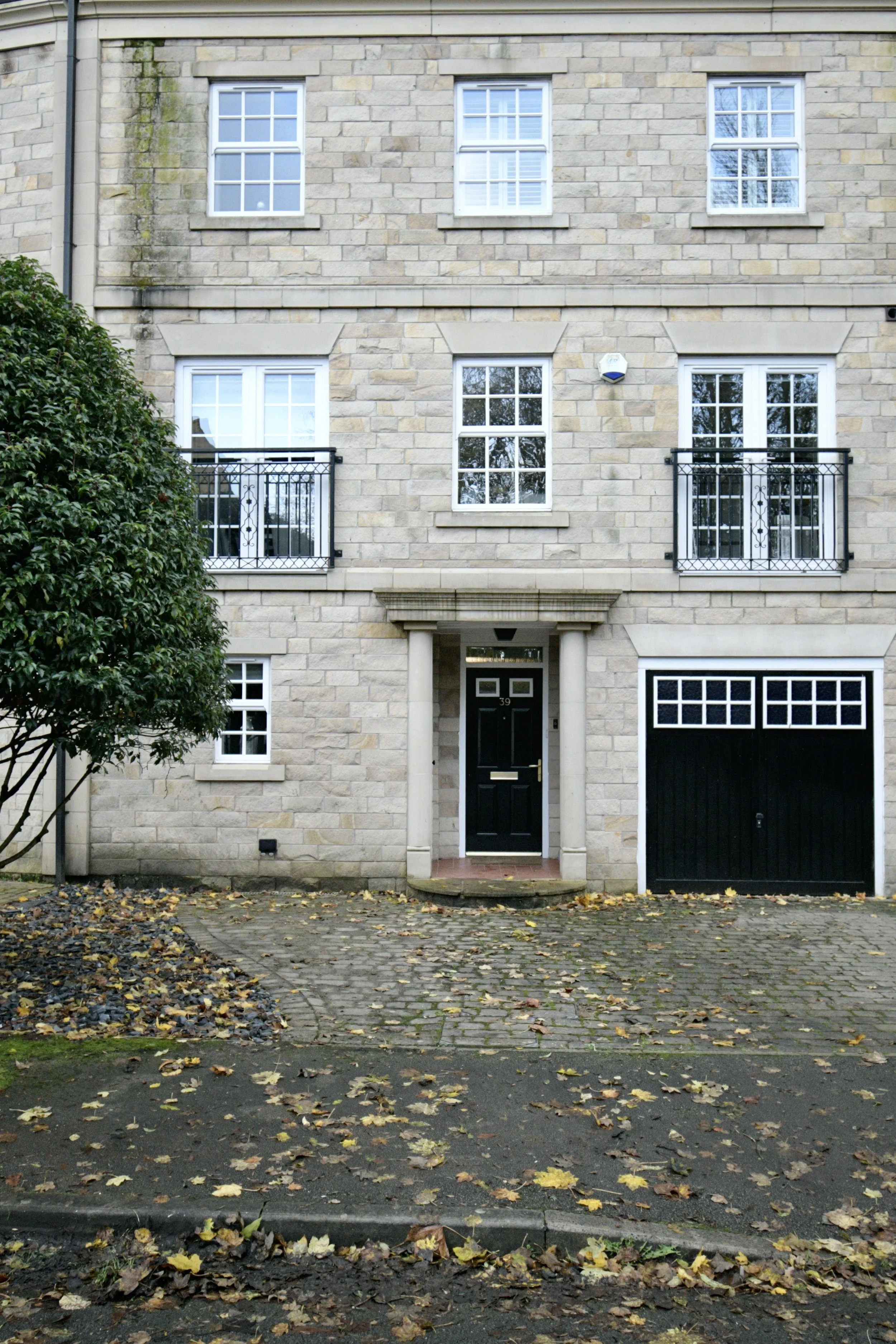 Front view of a multi-story beige brick house with white window frames and black garage door, surrounded by fallen autumn leaves and a green tree on the left.