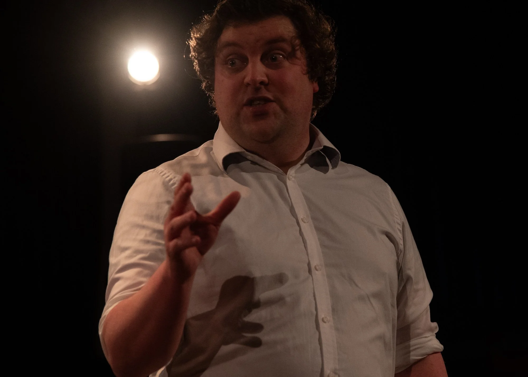 Drama scene: A man with curly hair wearing a white shirt speaking forcefully to us.
