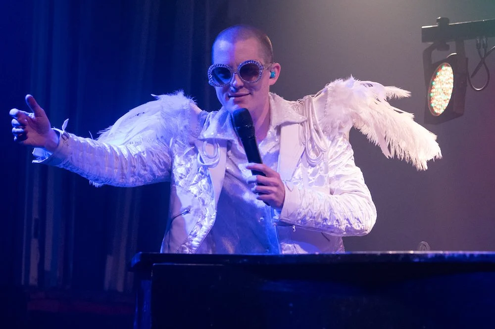 Elton John tribute dressed in an ornate white jacket with feathered shoulder accents and large sunglasses, holding a microphone on stage with colourful lighting.