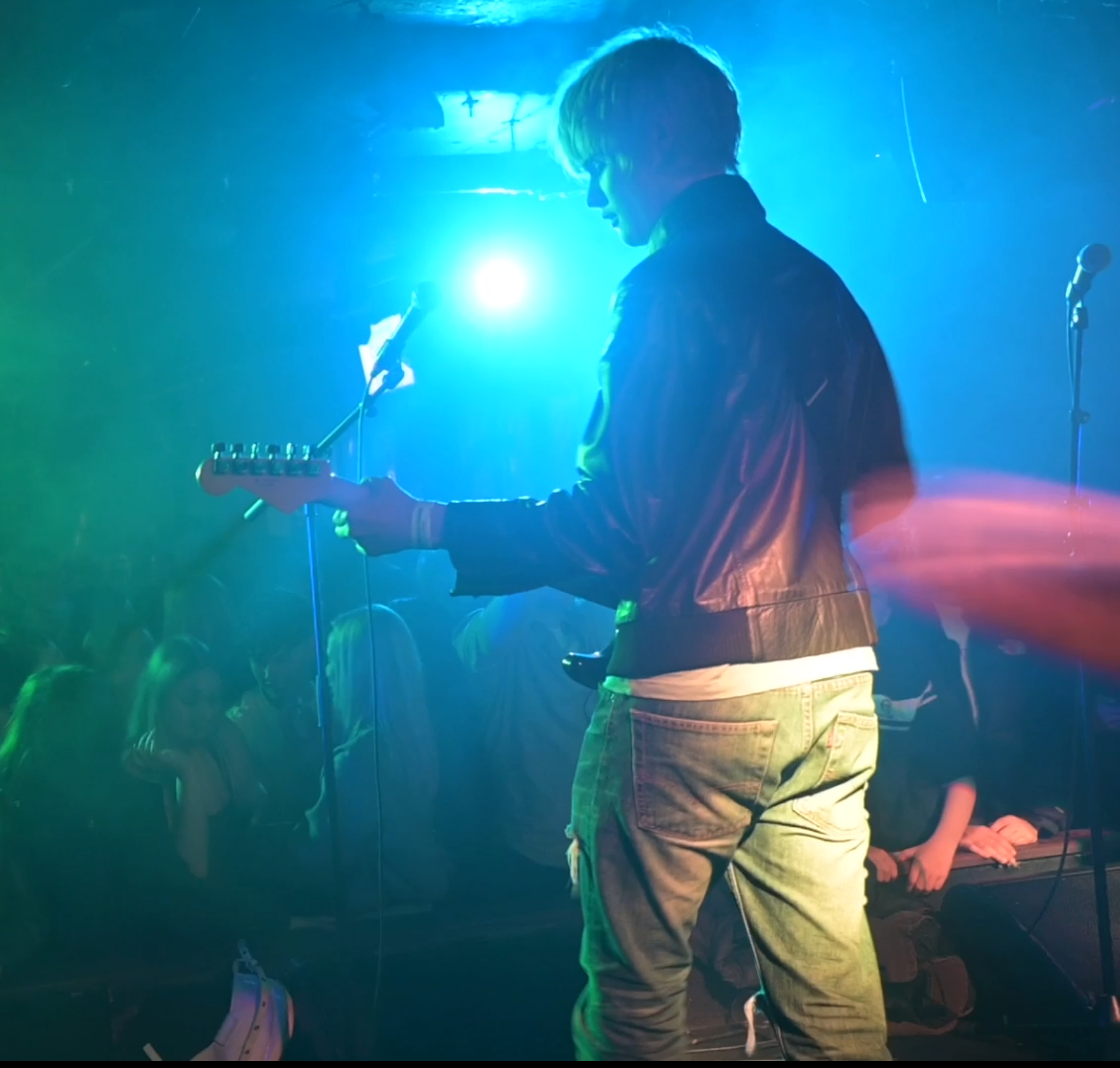 A guitarist wearing a dark jacket and light-colored jeans standing on a stage with vibrant blue and green lighting, with a crowd visible in the background.