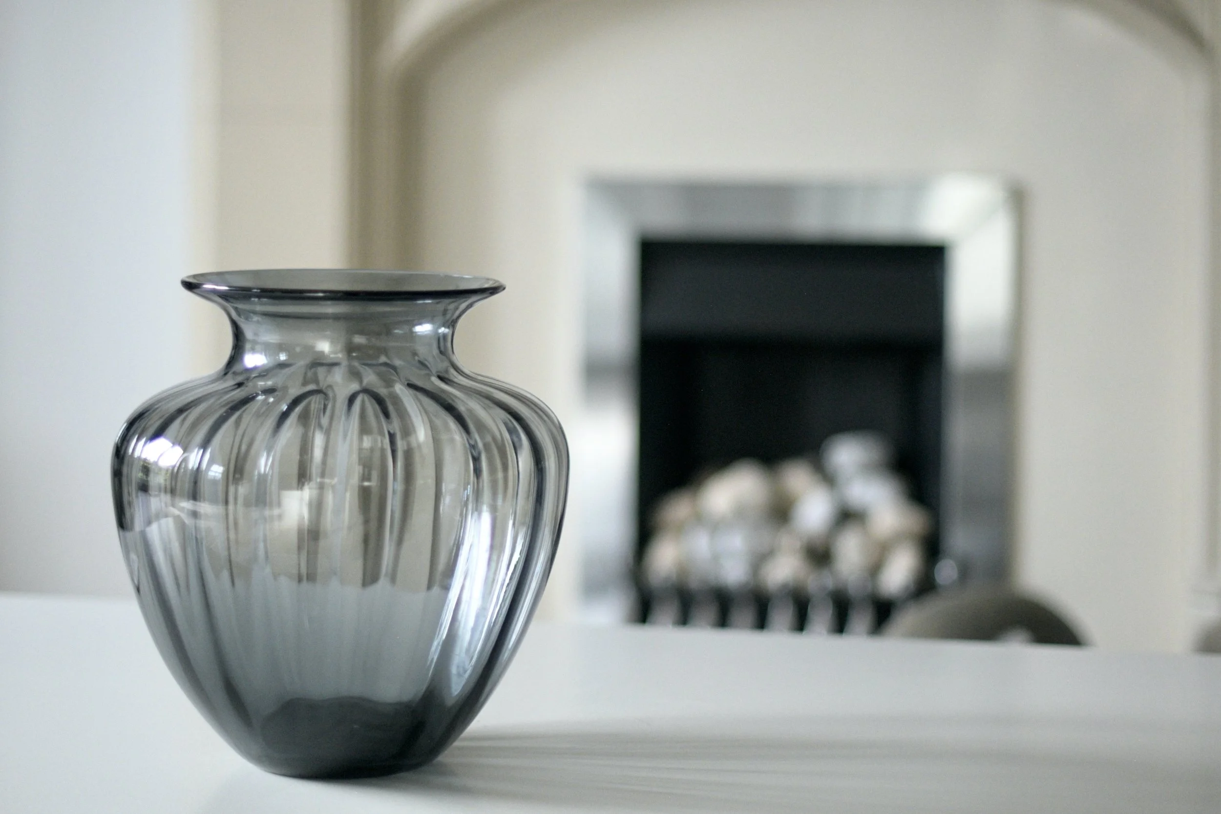 A clear, ribbed glass vase with a black rim on a white surface in front of a mirror or picture frame.