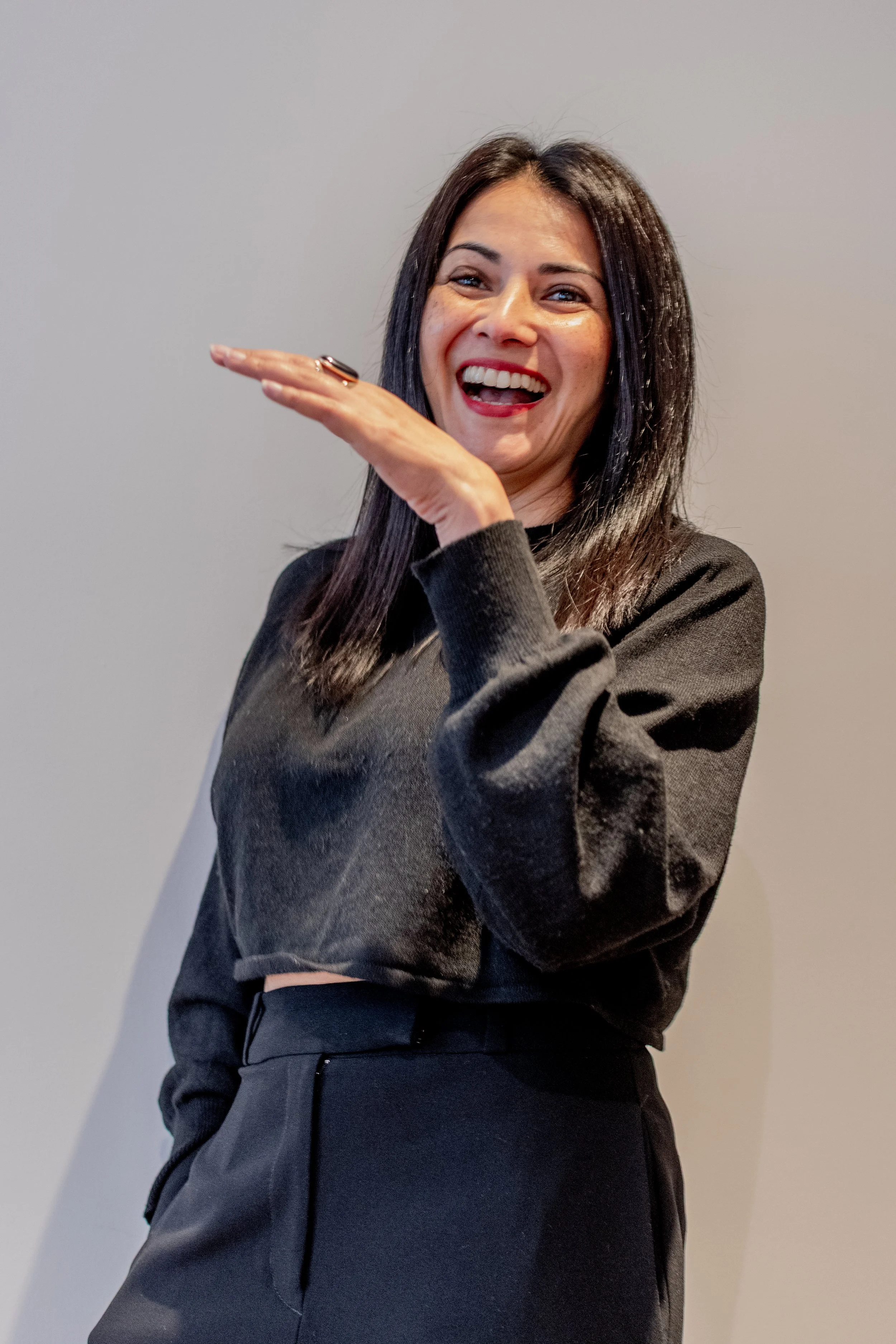 A woman with dark hair laughing and gesturing with her hand against a plain light-colored wall.