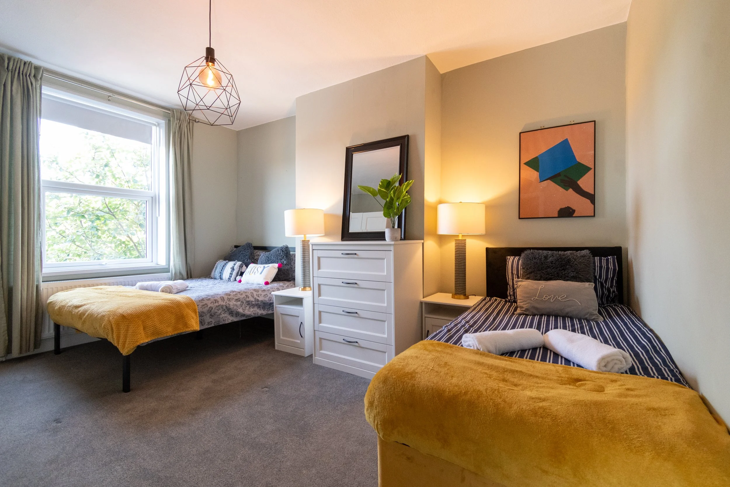 A bedroom with two beds, one with yellow bedding and the other with gray and white bedding, near a window with curtains. There are two nightstands with table lamps, a framed mirror, a potted plant, and wall art in the room.