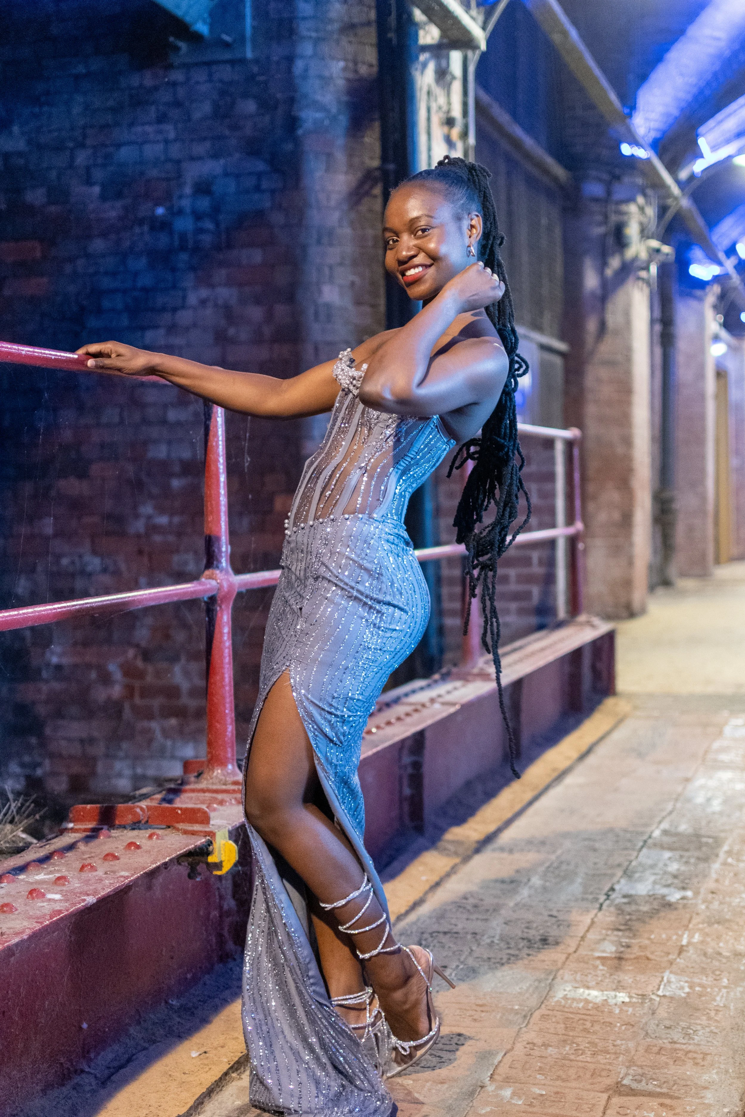 A woman in a silver, form-fitting, sequined dress with side slit. She has long, black dreadlocks and is wearing silver heels and jewelry. She is smiling, leaning against a pink railing with her left hand, and touching her neck with her right hand. The background includes a brick wall and urban lighting.