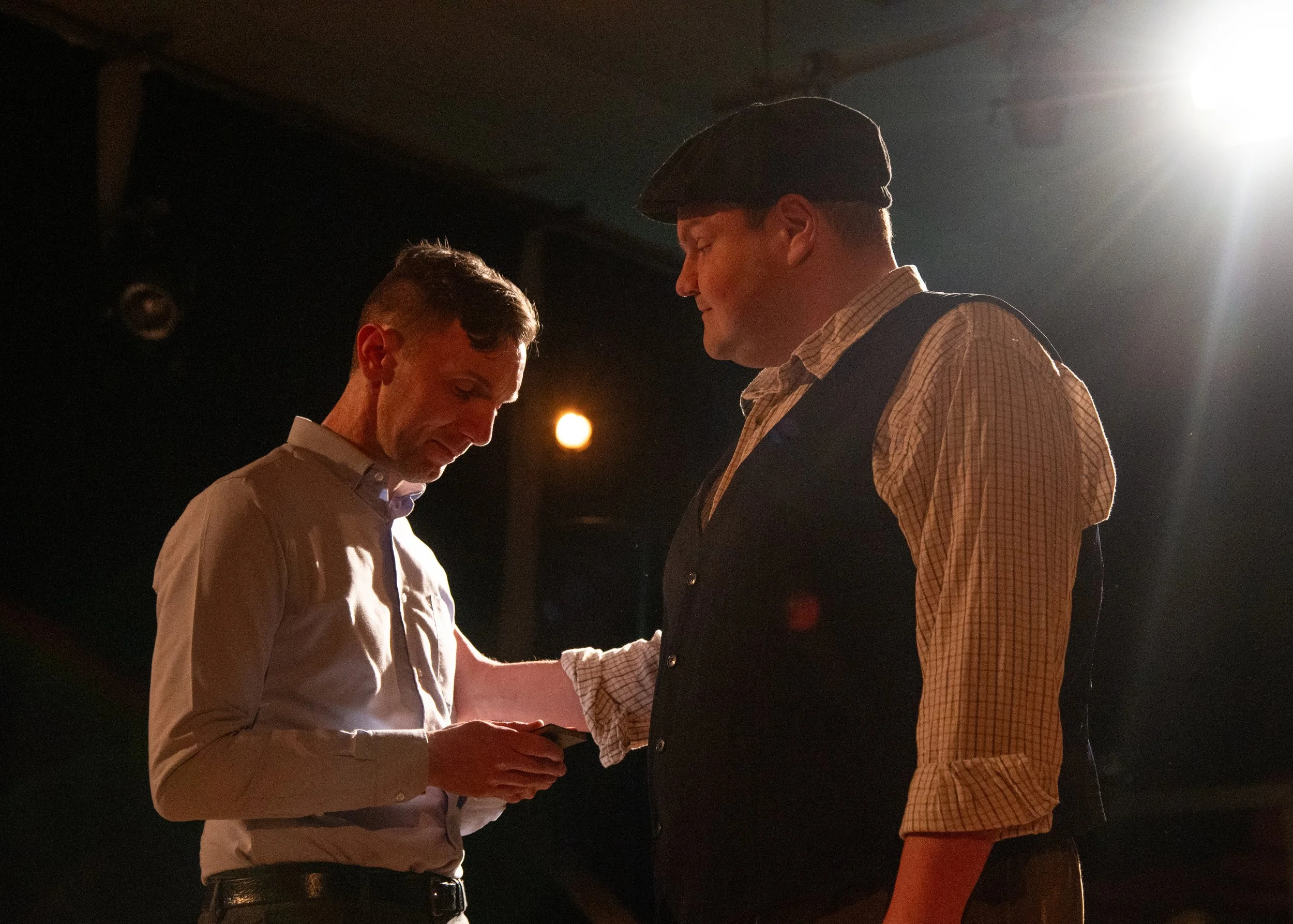 Drama scene: Two men in a dimly lit room, one with a beret and checkered shirt, the other with a collared shirt, sharing an emotional moment.