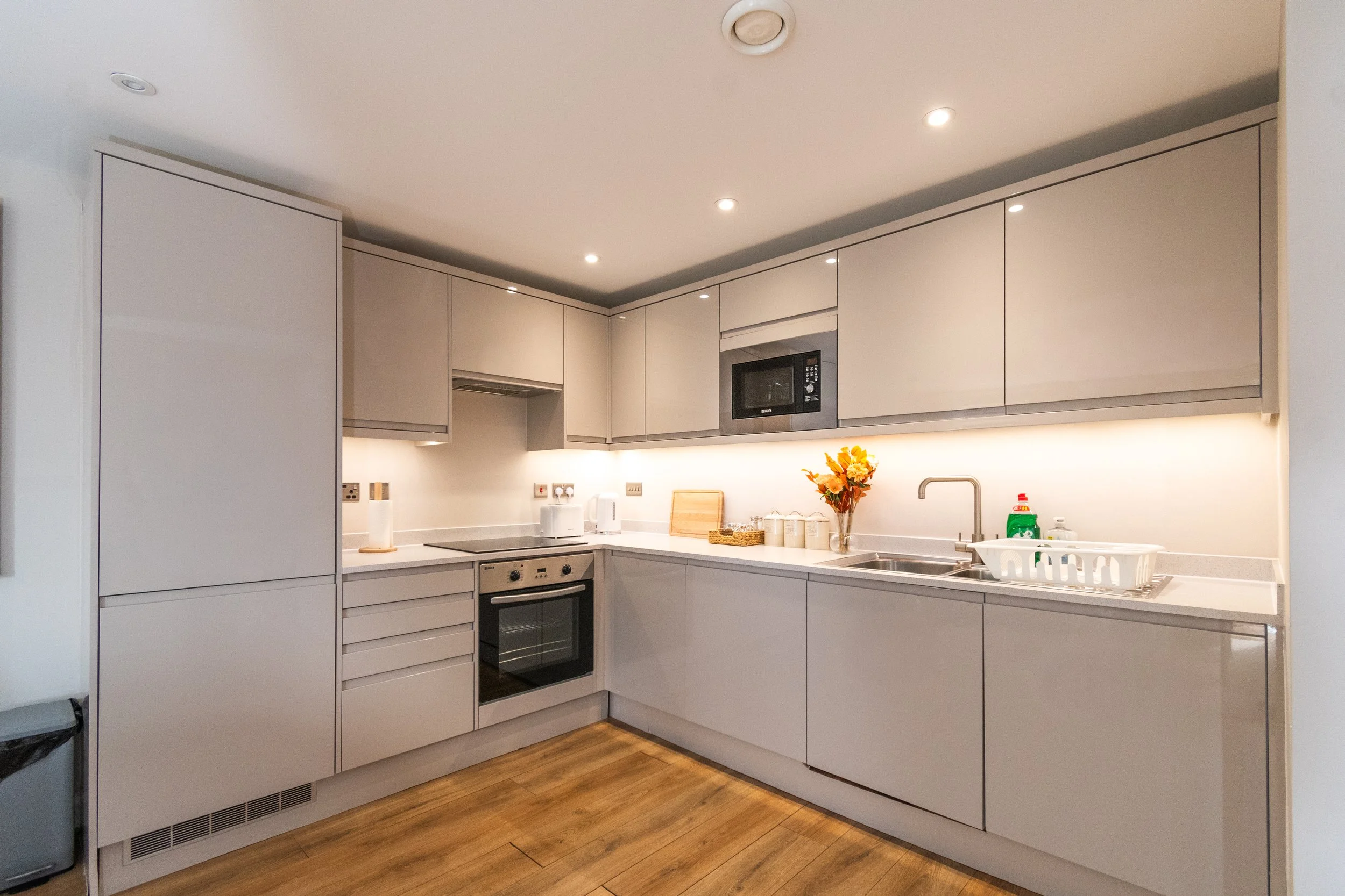 Modern kitchen with white cabinets, stainless steel appliances, wooden flooring, and a vase of orange flowers on the counter.