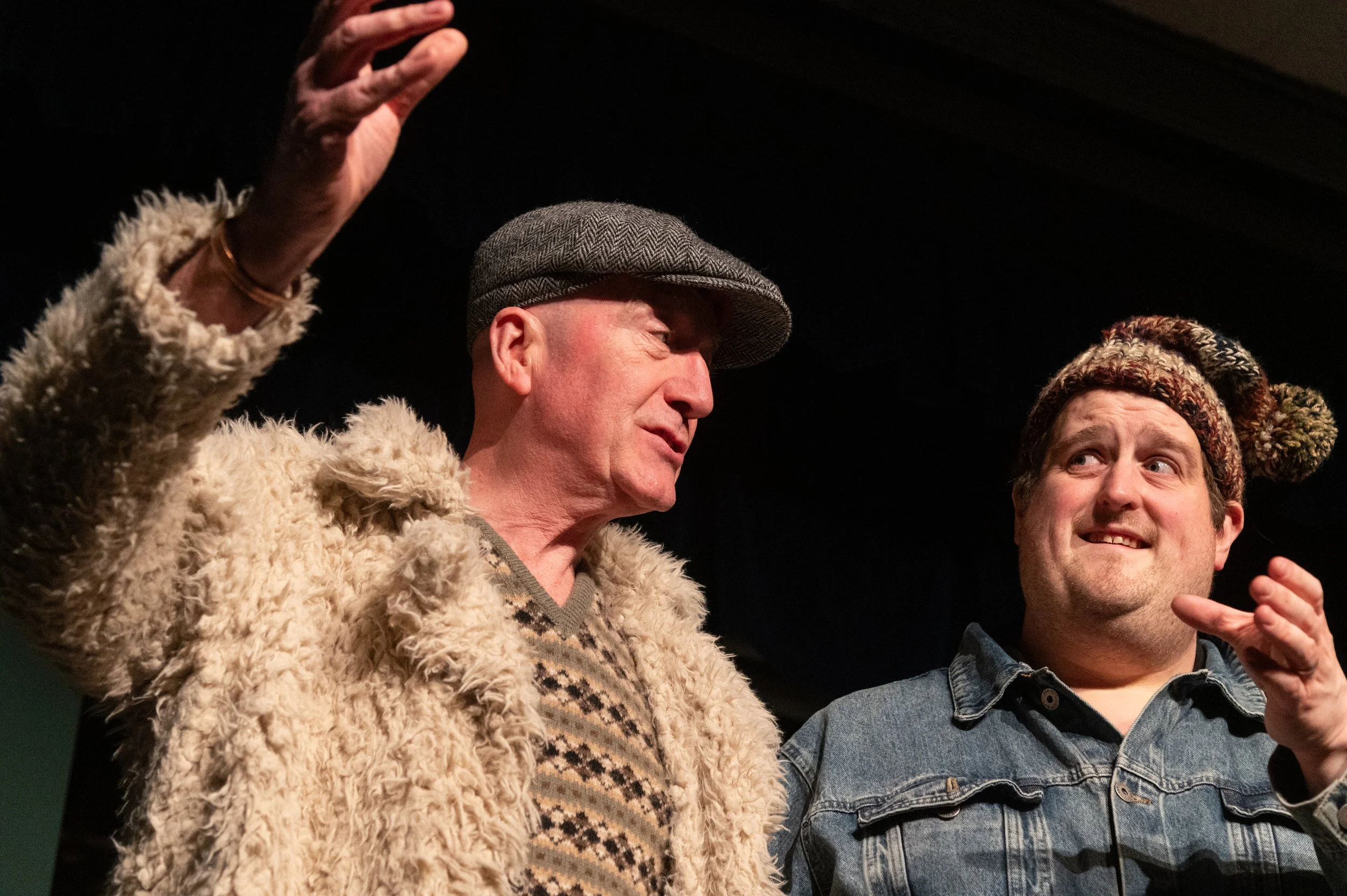 Two men engaged in humorous conversation, one wearing a gray herringbone flat cap and a beige shaggy coat, the other wearing a knitted hat with a pom-pom and a denim jacket, on a dark stage.
