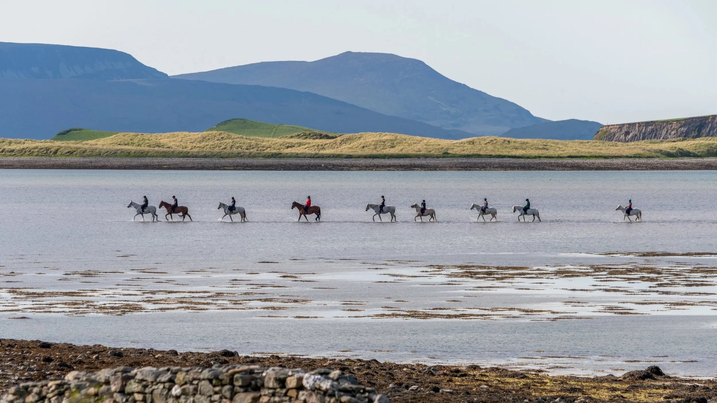 A group of people riding horses across a body of water with mountain hills in the background.