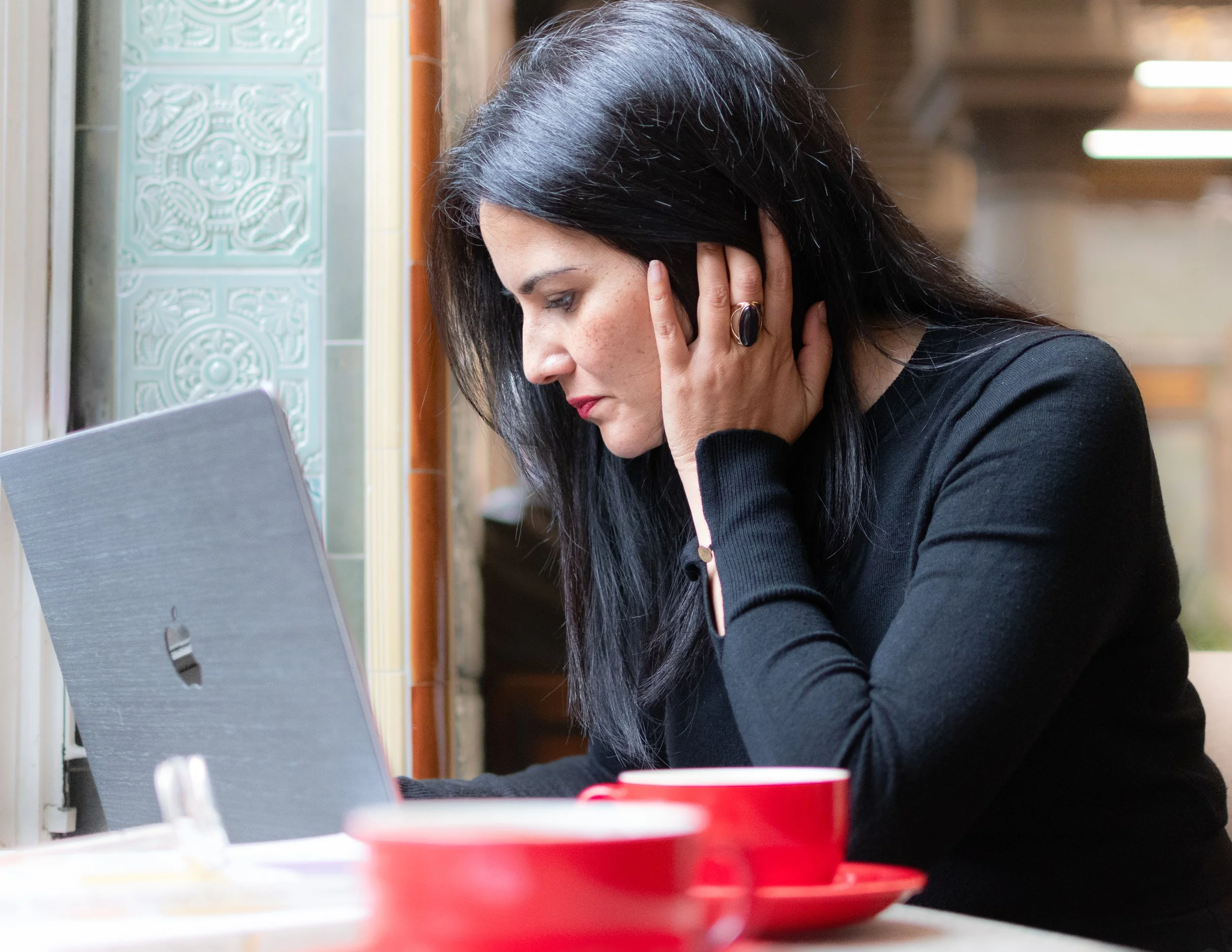 A woman with black hair and red lipstick, wearing a black long-sleeve shirt, is sitting at a table, resting her head on her hand, and studying her laptop, lit by the window in front of her.