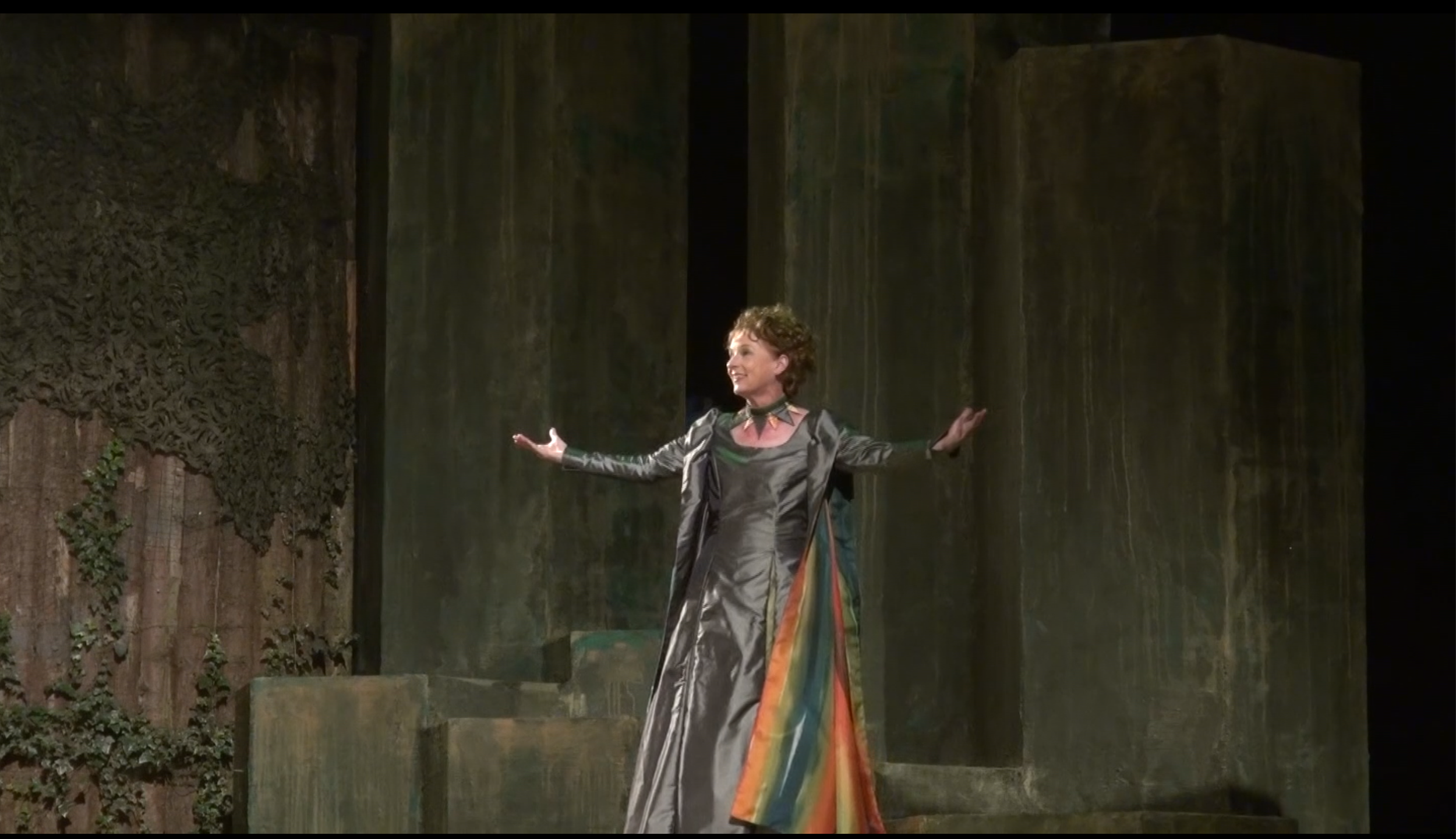 A woman dressed in a multicolored, flowing gown stands on a stage with arms outstretched, smiling. The background consists of large, textured, dark green and brown panels with some ivy growing along the sides.