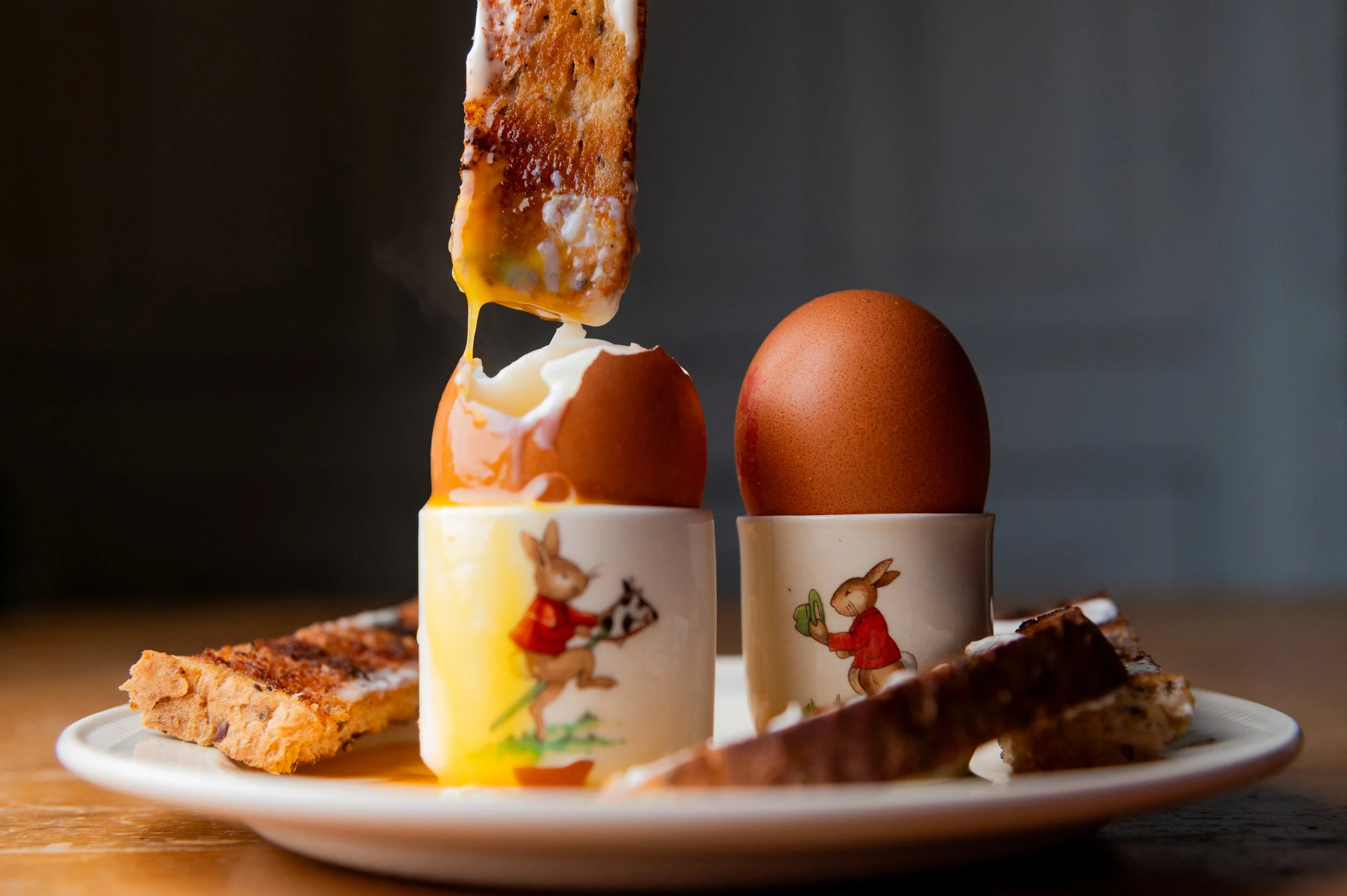 Toast soldiers and eggs in decorated egg cups on a white plate with dark background.