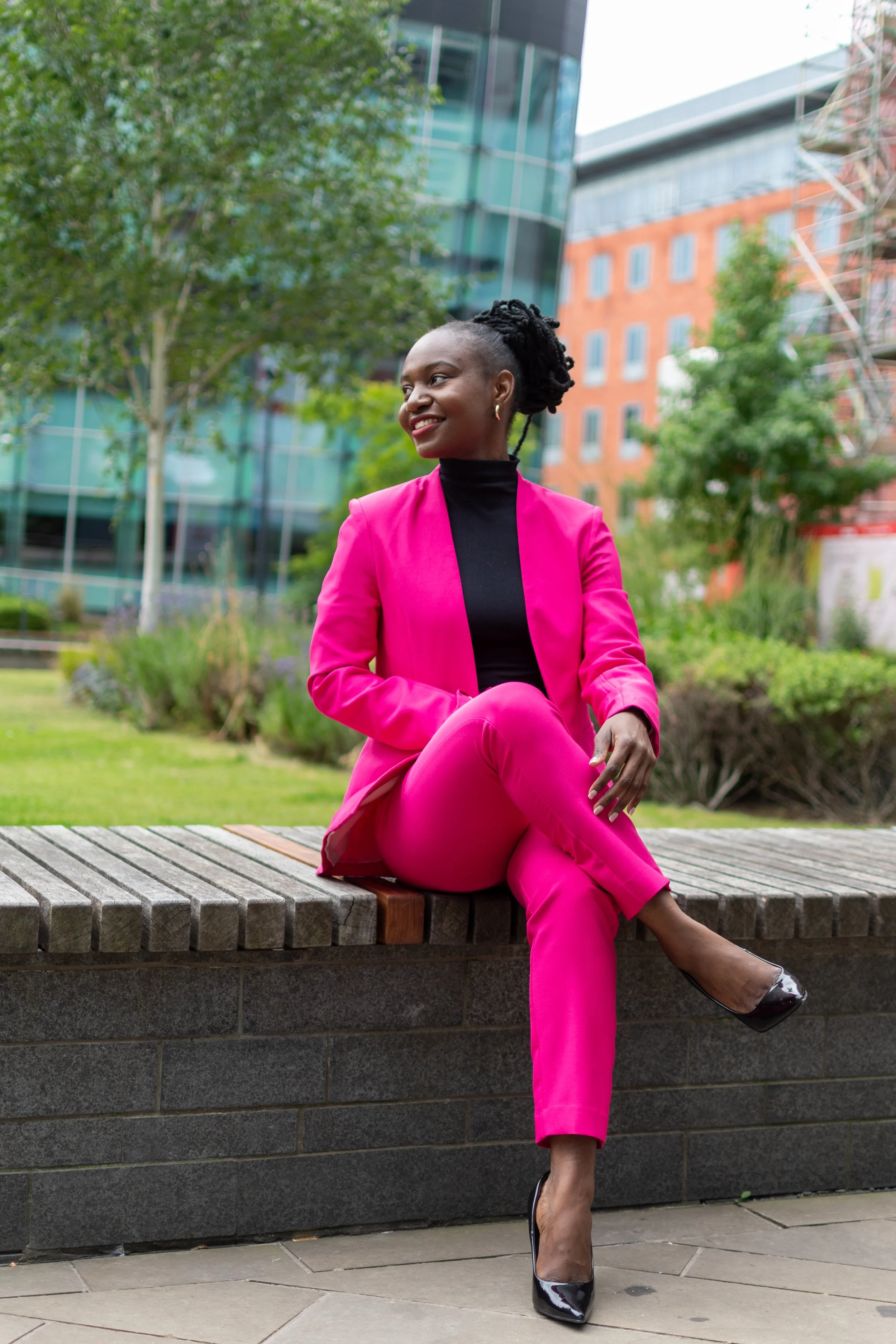 A woman in a pink suit and black heels sitting on a concrete barrier outdoors, smiling and looking to the side, with modern glass buildings and green trees in the background.