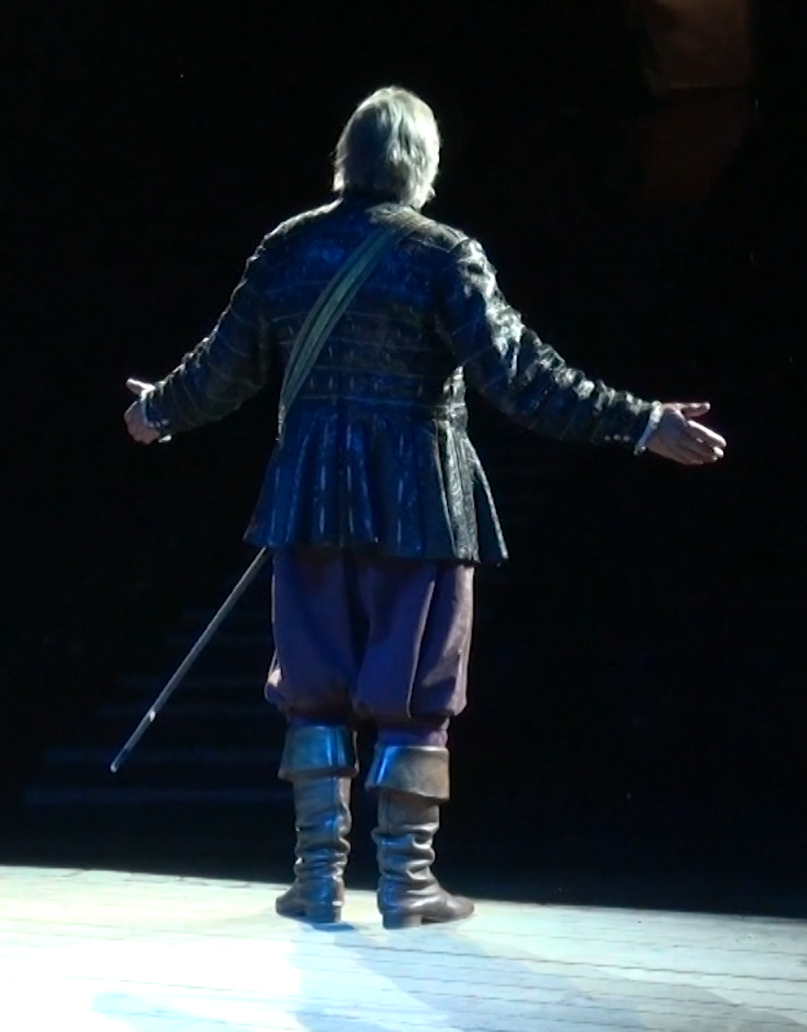 A performer dressed in period leather clothing, standing on stage with arms open, facing away from the camera, under spotlight against a dark background.