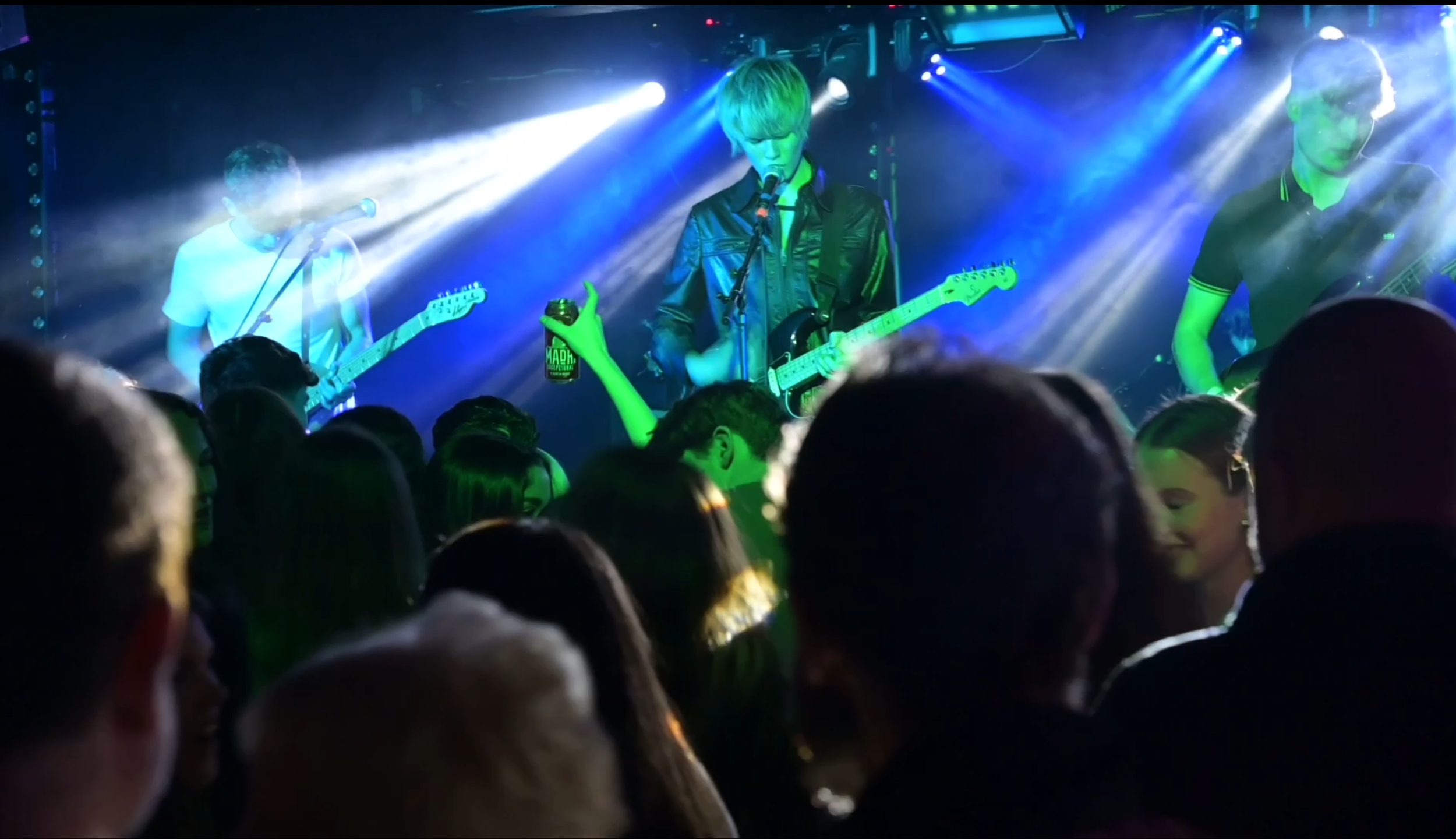 A band performing live on stage with three members playing guitars and singing, colorful stage lighting, and a crowd watching.
