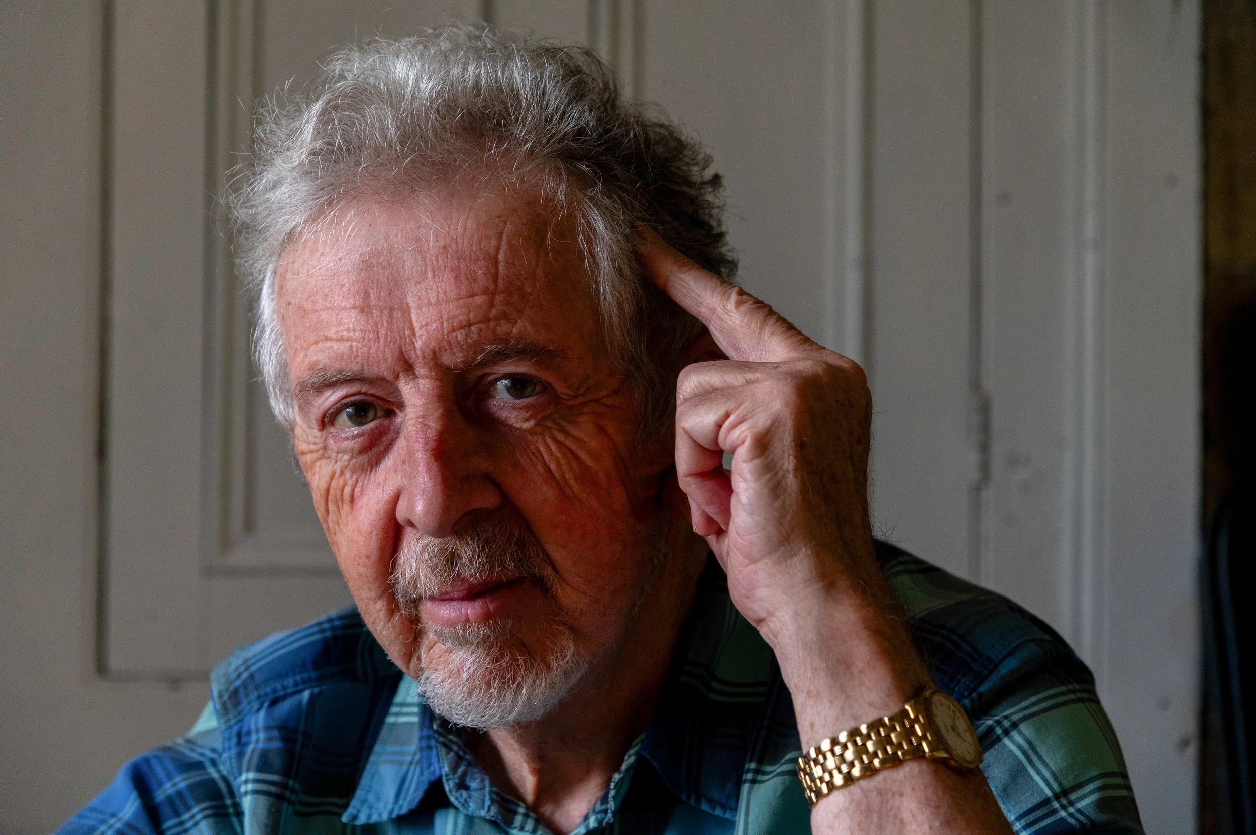 Close-up of an older man with grey hair and a beard, touching his temple with his right index finger, wearing a gold wristwatch and a blue plaid shirt, standing in front of a wooden wall.
