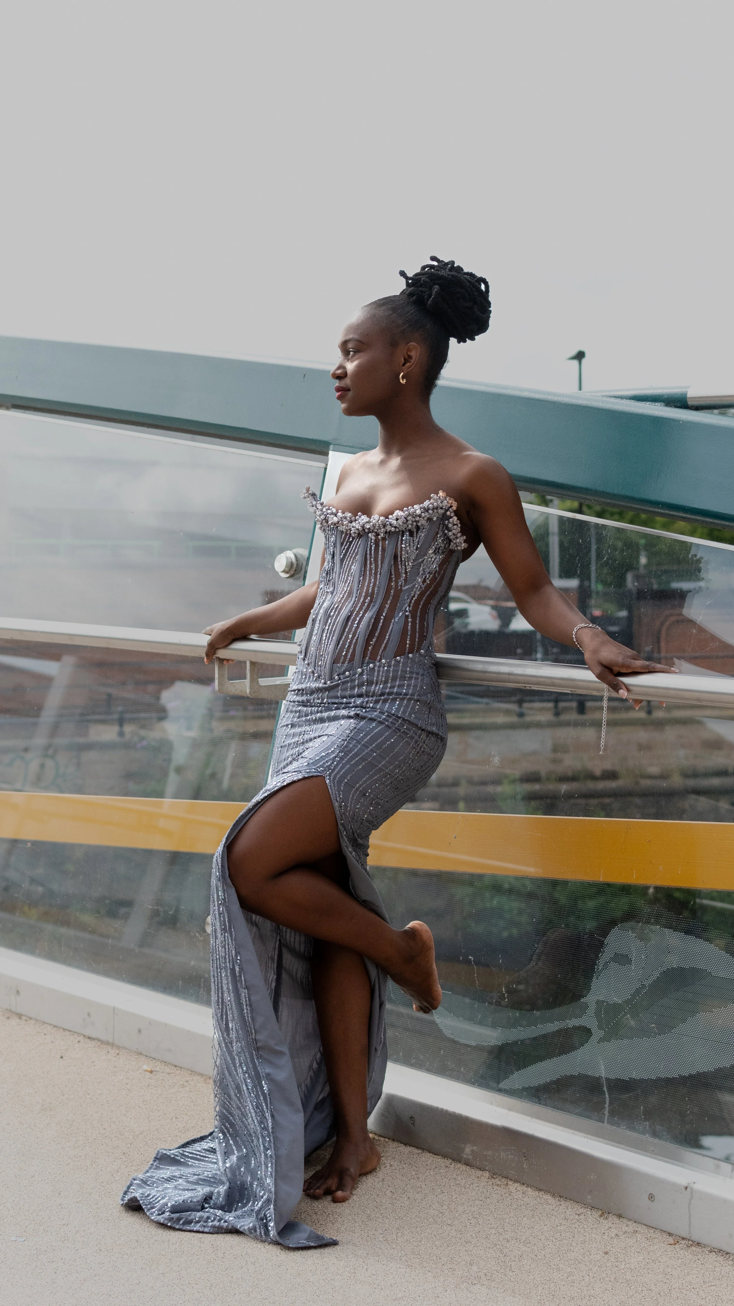 An elegant woman in a silver, sequined evening gown with a high slit, relaxing in daylight, at an outdoor location, with her eyes closed and legs crossed.