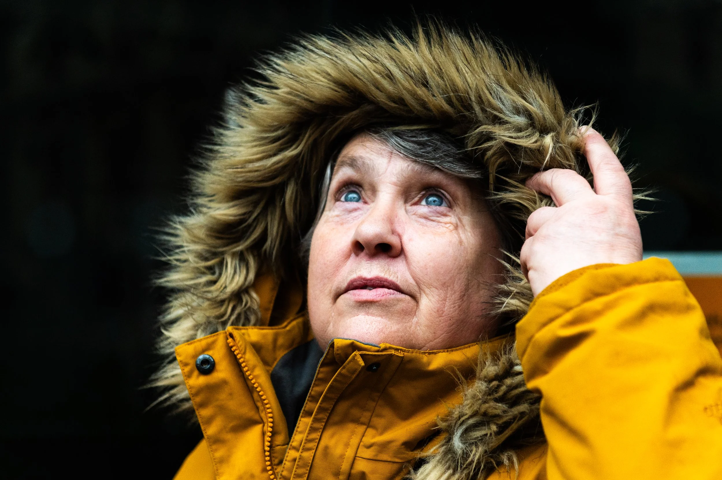 A woman with blue eyes gazing skywards, pulling back the hood of her yellow fur-lined jacket.
