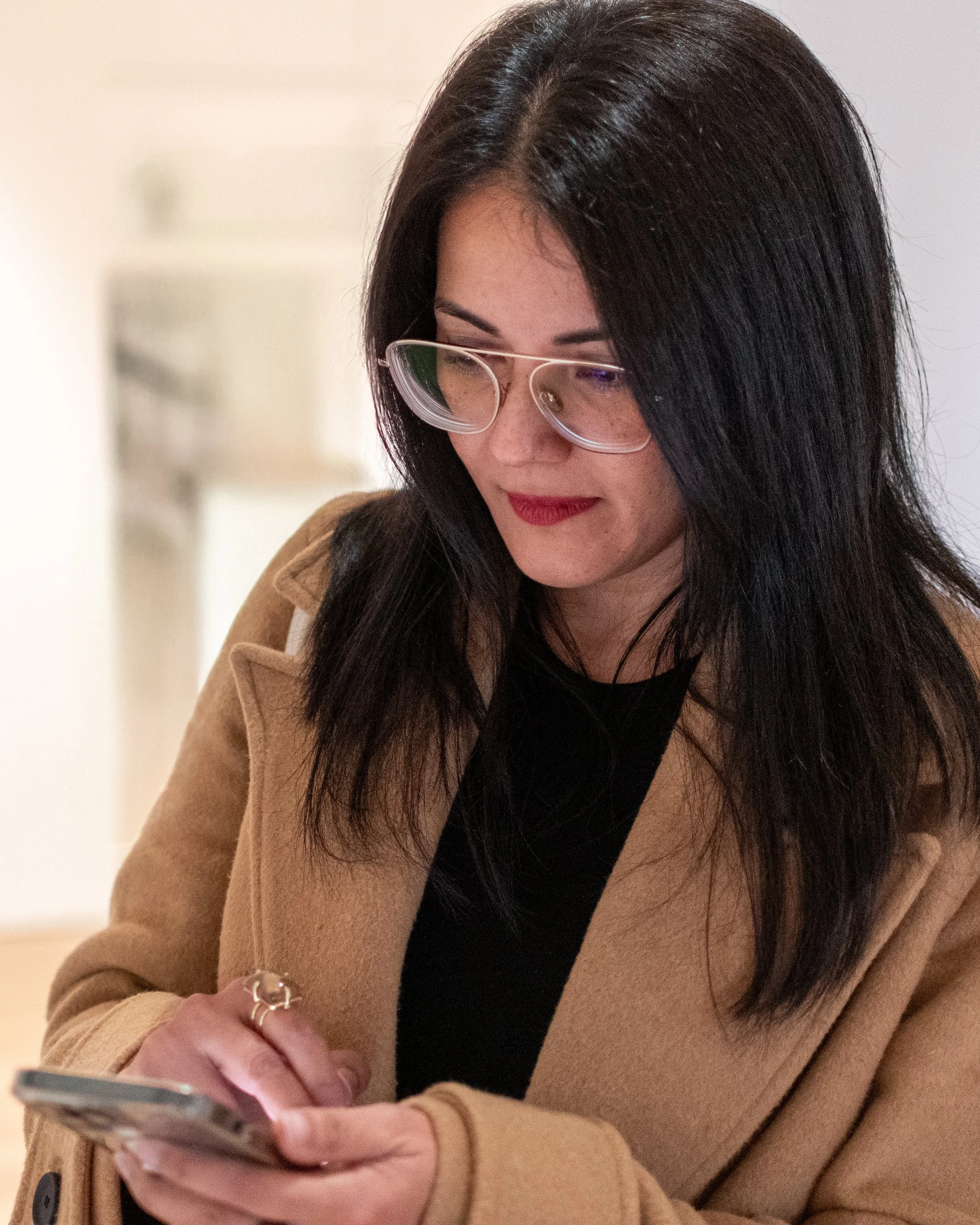 A woman with long dark hair and glasses looking down at her phone, wearing a tan coat over a black top.
