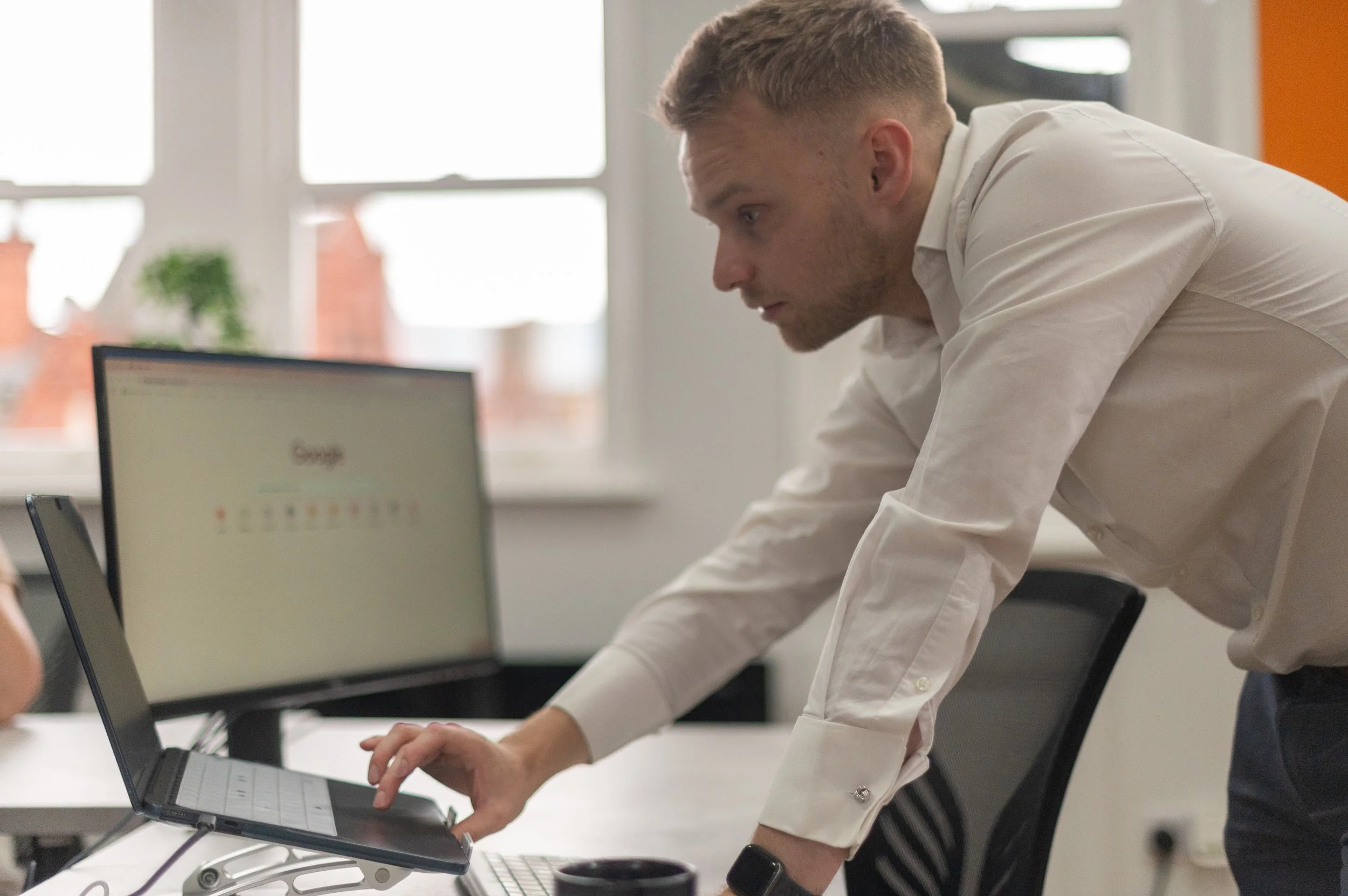 A young man with short hair and a beard leaning over a desk, working on a laptop in an office with large windows and a potted plant in the background.