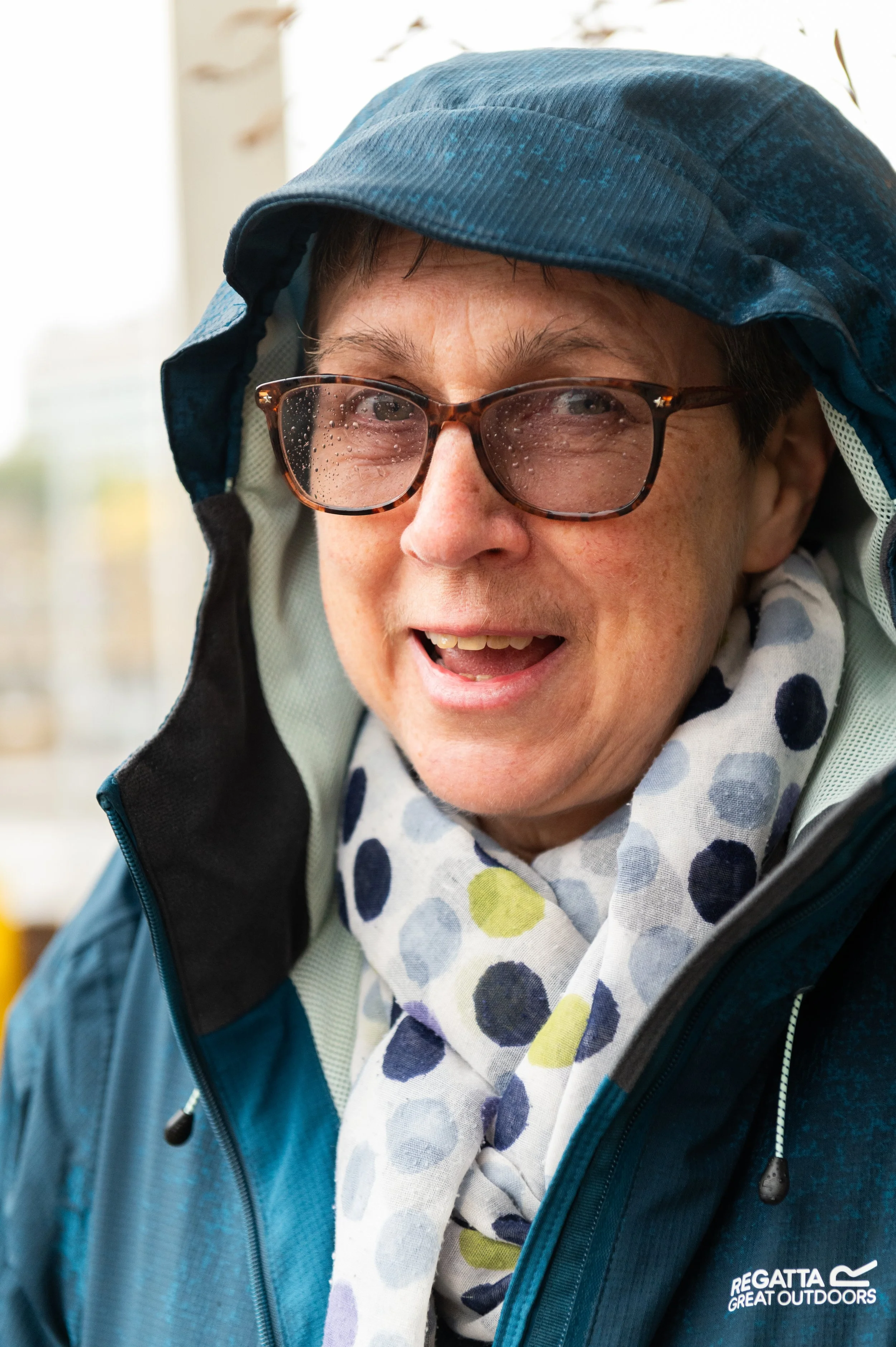 Close-up of a smiling person wearing glasses, a rain hood, and a jacket with a logo that says "Regatta Great Outdoors". They are also wearing a scarf with large colorful polka dots.
