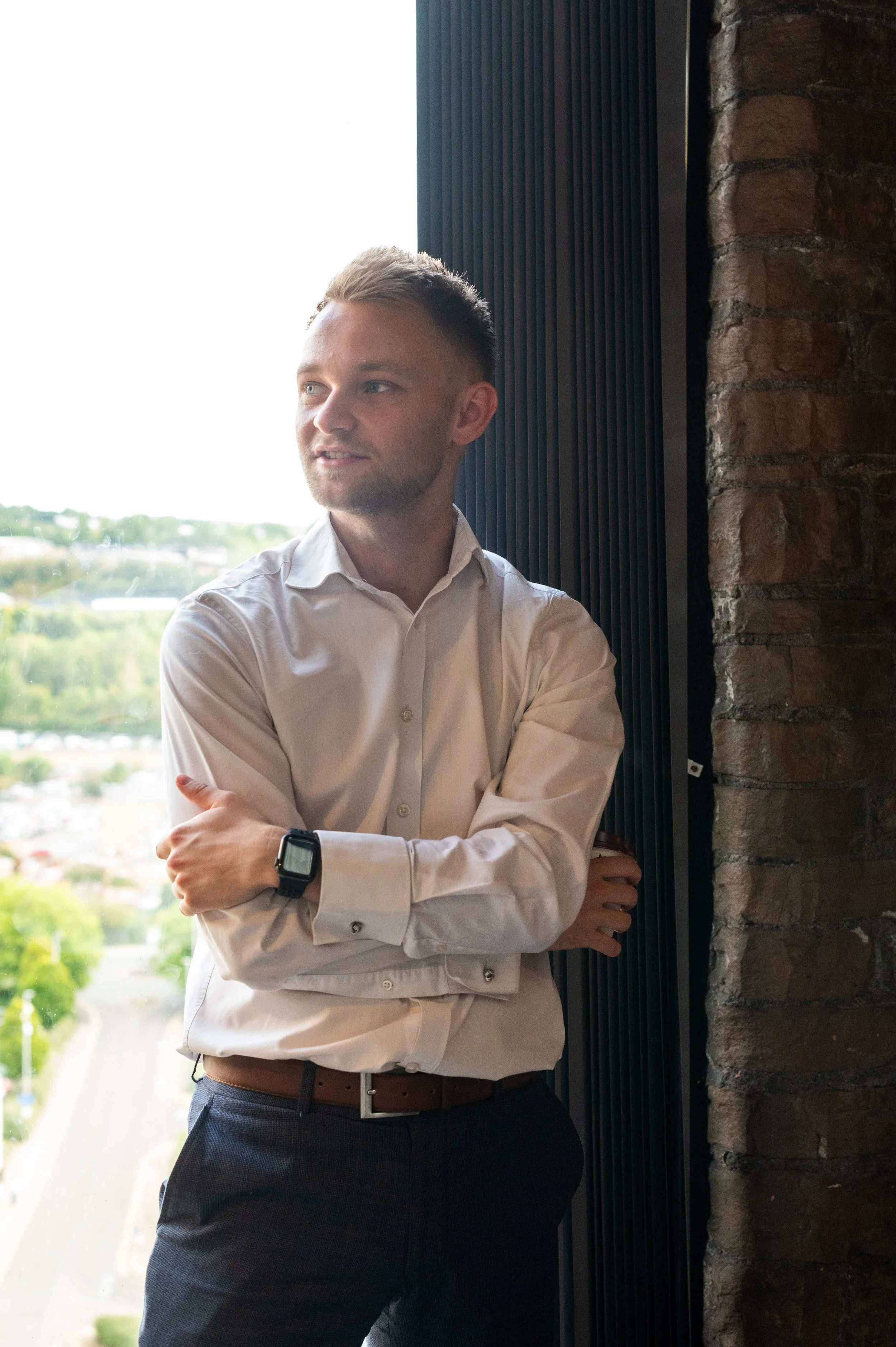 A young man with short blond hair and a beard, wearing a white dress shirt and dark pants, standing indoors near a brick wall and a large window with a view of a green landscape outside.