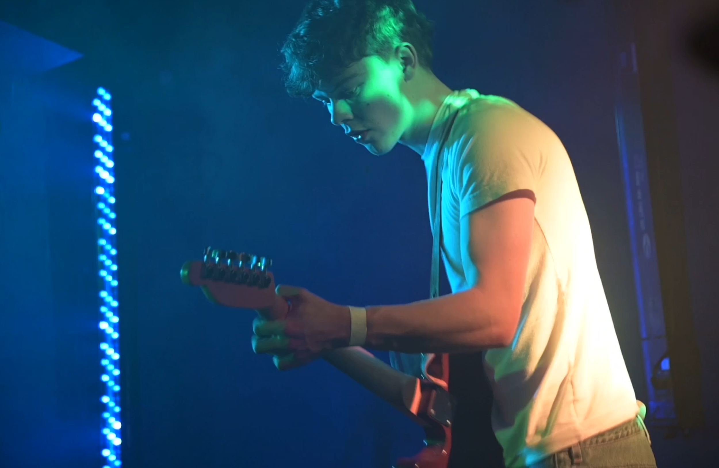 A guitarist with short curly hair playing onstage with blue and green lighting.