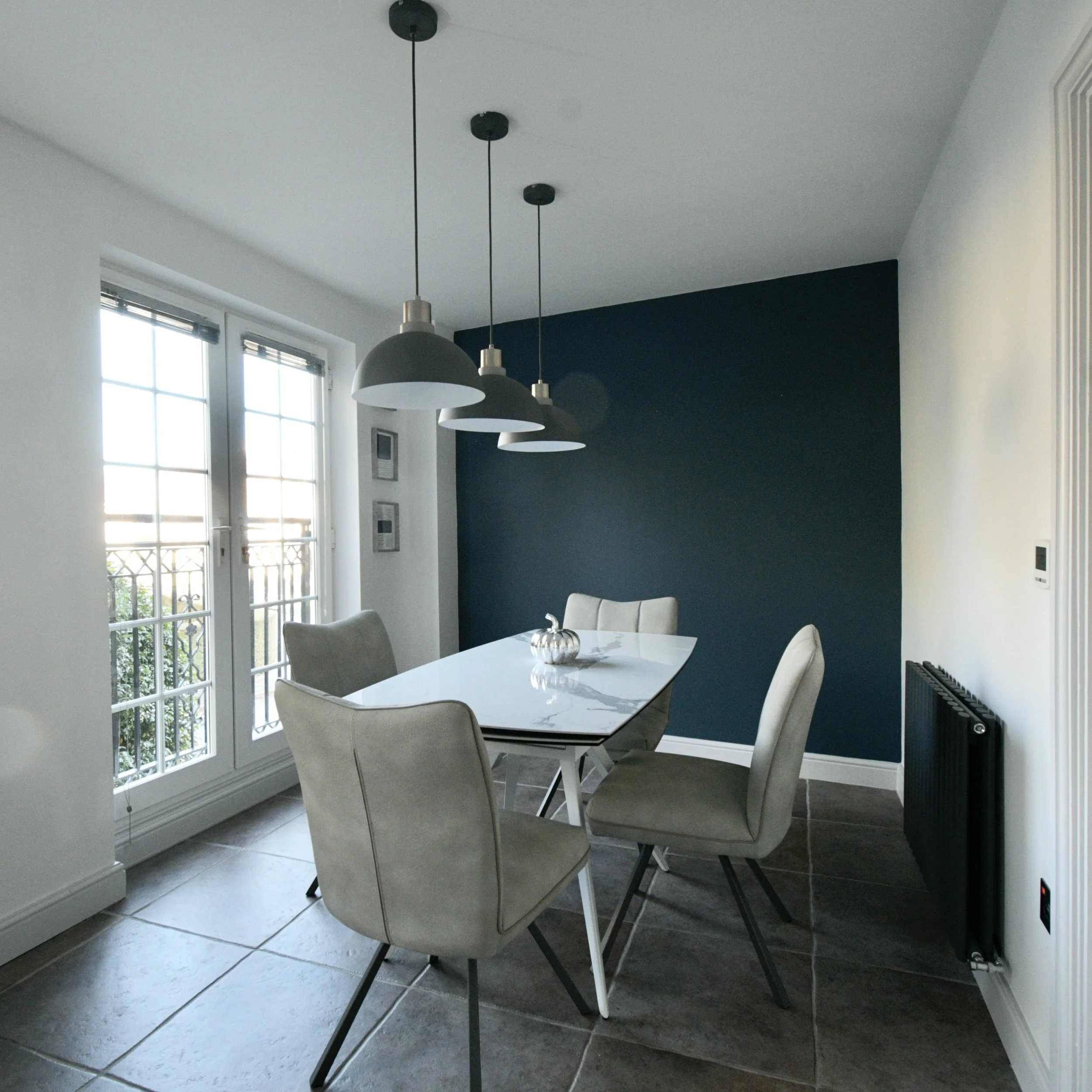 A dining area with a white table and four beige chairs, three pendant lights hanging from the ceiling, large windows with blinds letting in natural light, dark flooring, and a dark accent wall on the right side.