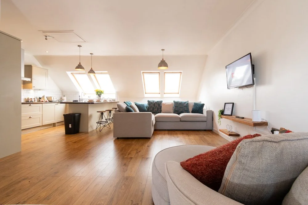 Living room with a grey sofa and a beige armchair, wooden flooring, and a wall-mounted TV, in an attic space with two skylight windows and a kitchen area in the background.