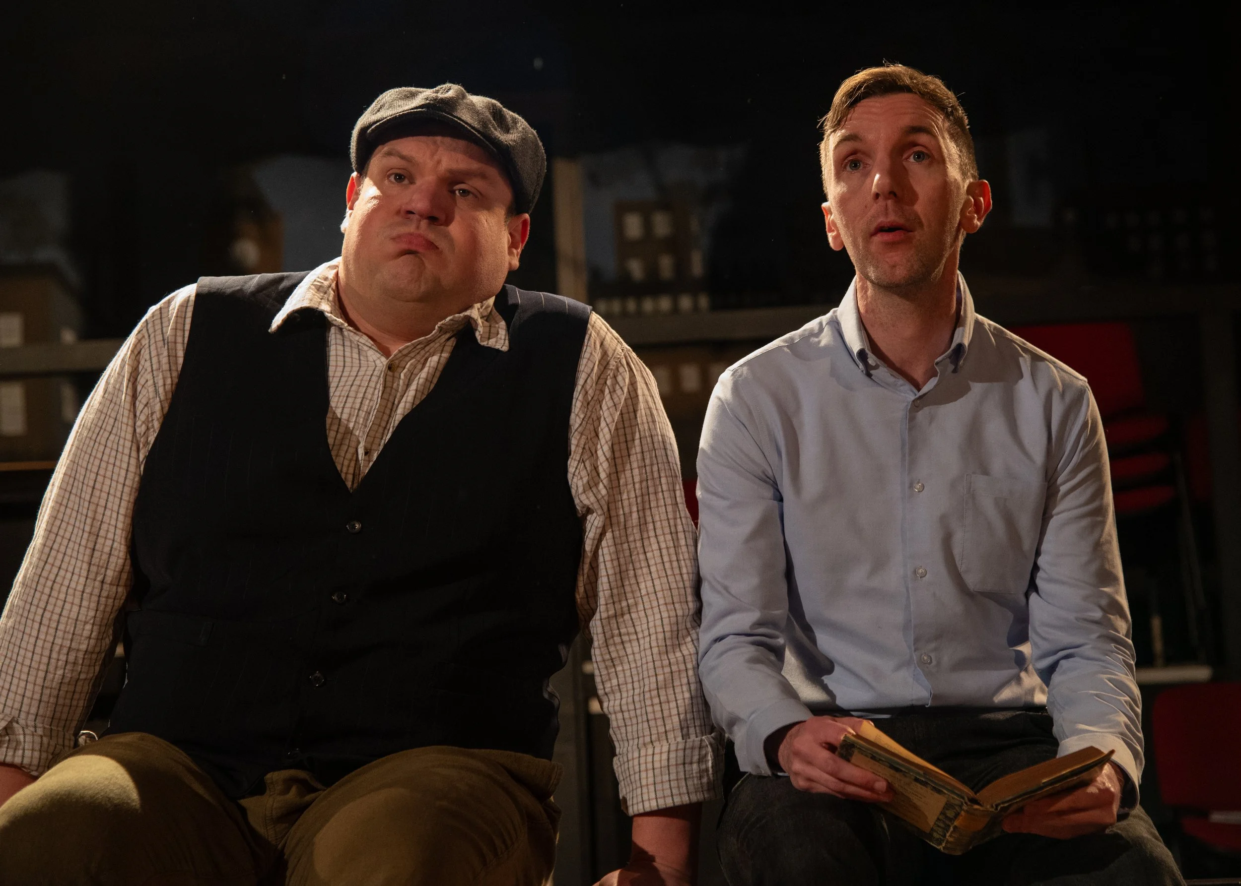 Drama scene: Two men sitting on a bench, one wearing a flat cap and a vest, the other holding a book, in a dimly lit indoor setting.