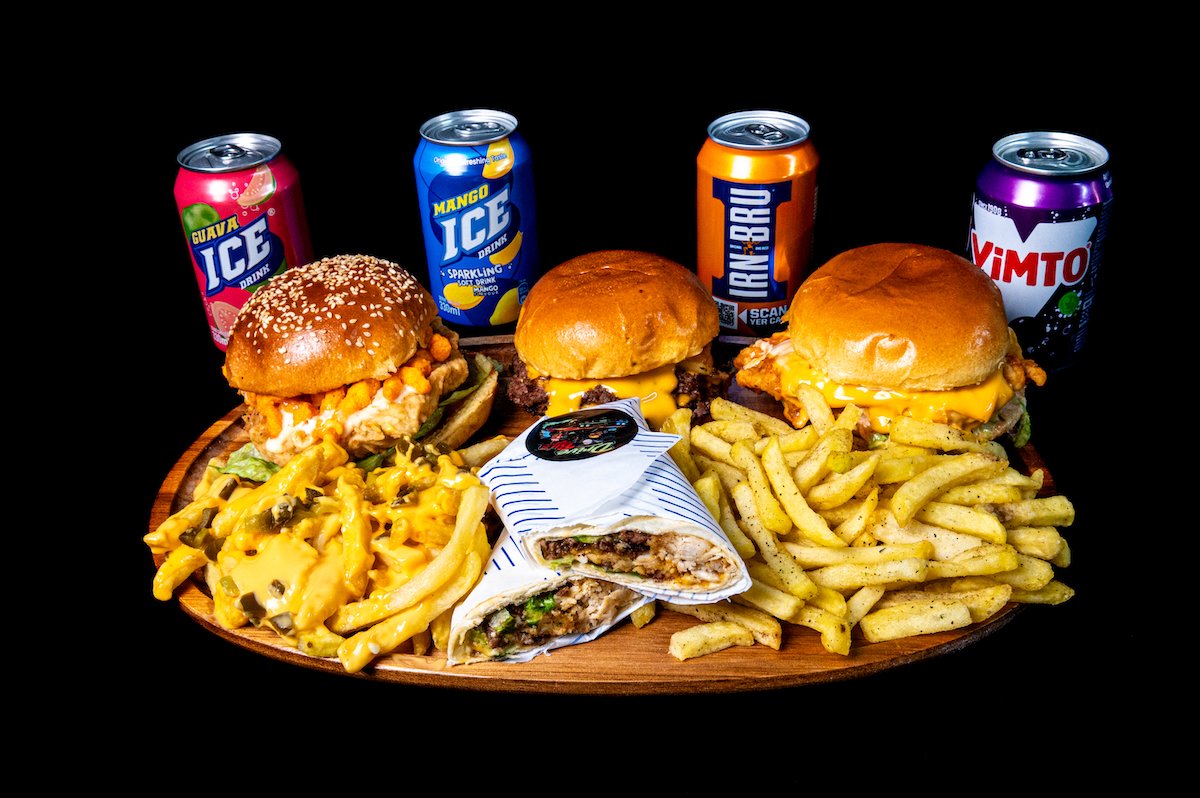 A wooden platter with three cheeseburgers, a serving of French fries, a wrap, and three cans of soft drinks against a black background.